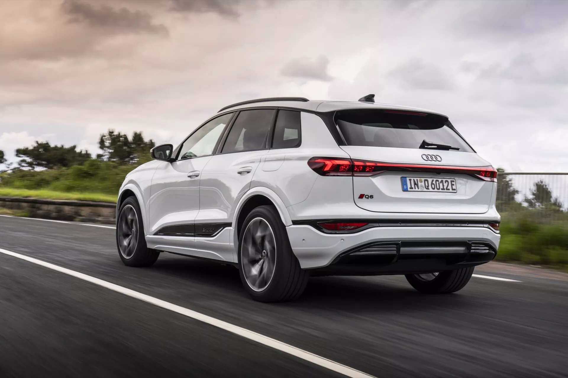 Audi Q4 e-tron 2024 jadący po asfaltowej drodze, widziany z trzy czwarte tylnego widoku, ukazujący lewą tylną stronę. Zdjęcie pokazuje smukłe tylne światła, spojler dachowy, panoramiczny dach, wieloramienne felgi aluminiowe, dwukolorowe nadwozie z metalicznym obramowaniem i ikoniczny znak Audi. Rozmycie ruchu kół i zieleń przy drodze z pochmurnym niebem sugerują dynamiczne warunki jazdy w każdym pogodzie.