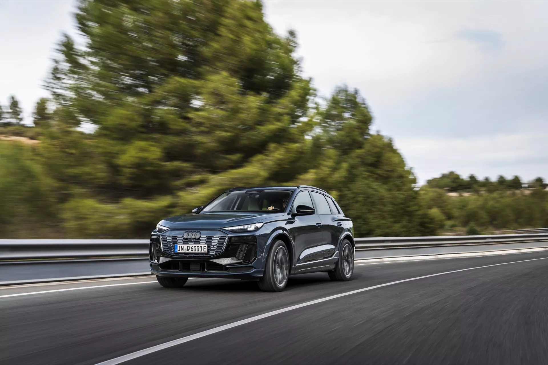 Audi Q6 e-tron 2024 SUV w ruchu na autostradzie, uchwycony z przodu w trzech czwartych. Obraz pokazuje przedni grill, reflektory LED, maskę, przednią szybę i część bocznego profilu, w tym drzwi i lusterka boczne. Pojazd jedzie po prawej stronie pasa, poruszając się z lewej do prawej z rozmazanym tłem wskazującym na prędkość. Drzewa i barierka są nieco widoczne. Czysty design przednich reflektorów sugeruje zaawansowane systemy oświetleniowe, a duży grill wskazuje na technologie chłodzenia lub czujniki dla ADAS. Lusterka boczne mogą zawierać wskaźniki kierunku lub systemy monitorowania martwego pola. Koła dostarczają wgląd w kategorie wydajności lub luksusu.