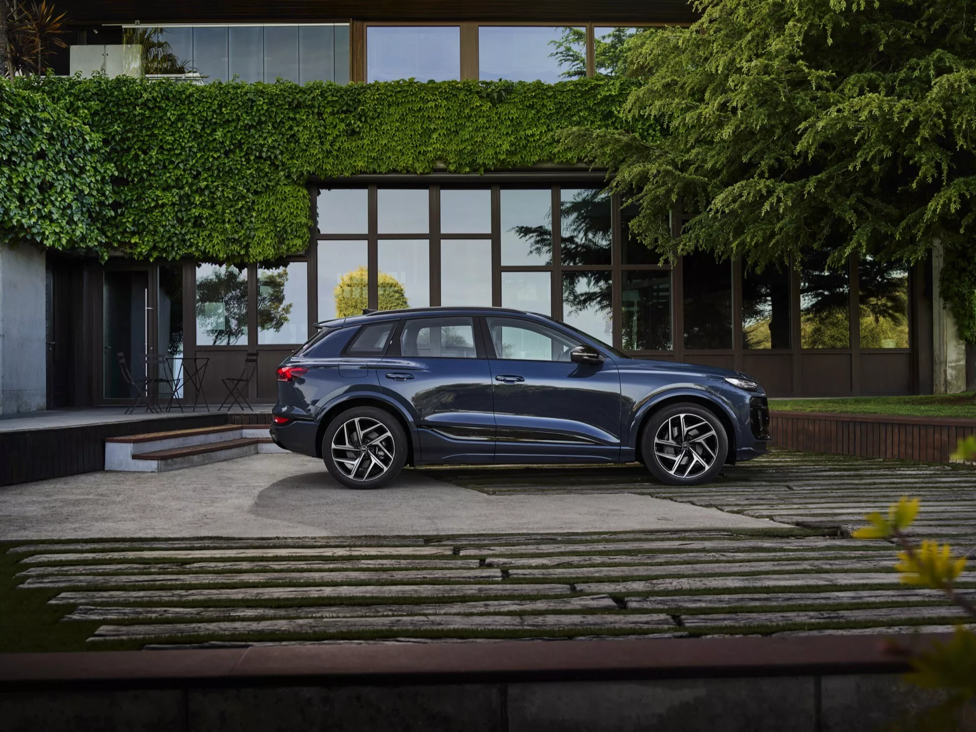 Audi Q6 e-tron 2024 stoi na dobrze zagospodarowanym podjeździe, uchwycona w pełnym widoku profilu z nieco podwyższonego kąta. Głęboki metaliczno-niebieski SUV błyszczy eleganckim designem, z aerodynamicznymi liniami dachu, przyciemnianymi szybami i równymi klamkami drzwi. Wieloszprychowe felgi aluminiowe i niskoprofilowe opony podkreślają jego luksusowe wykończenie. Na tle nowoczesnego luksusowego budynku z dużymi szklanymi oknami i kamienną i trawiastą podjazdem, scena jest delikatnie oświetlona, podkreślając premium status pojazdu.
