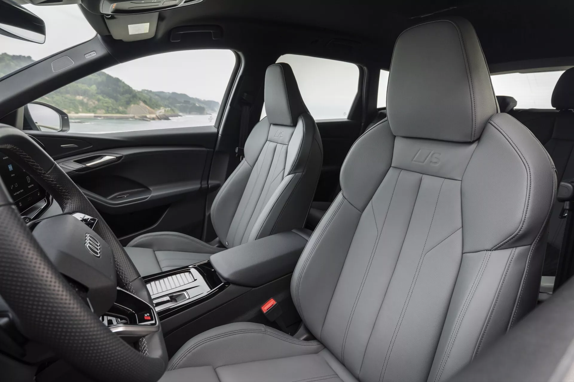 Widok wnętrza Audi Q6 e-Tron 2024 prezentujący luksusowe skórzane przednie fotele z wyraźnym wzorem szwów, wytłoczonym logo i nowoczesnym designem. Fotografia podkreśla deskę rozdzielczą z kierownicą z zamontowanymi przyciskami oraz nieoświetlonym cyfrowym panelem wskaźników. Częściowo widoczna jest konsola centralna ze stylową dźwignią zmiany biegów oraz starannie rozmieszczonymi przyciskami. Obraz ukazuje wyściełane podłokietniki drzwi z przyciskami do sterowania lusterkami, oknami i prawdopodobnie ustawieniami foteli. Światło naturalne podkreśla komfort i wysoką jakość materiałów we wnętrzu kabiny.