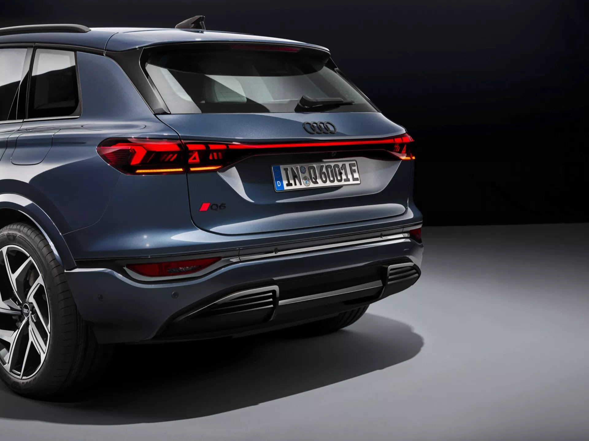Ten obraz przedstawia nowoczesny SUV, prawdopodobnie Audi Q6 e-tron 2024, widziany z podwyższonej tylnej perspektywy. Kultowe czteropierścieniowe logo Audi jest wyraźnie widoczne z tyłu, otoczone eleganckim ciągłym paskiem tylnych świateł LED, który rozciąga się na całą szerokość pojazdu, z segmentowymi światłami hamowania i kierunkowskazami. Samochód w ciemnym granatowym kolorze, z metalicznym połyskiem, odważnym wzorem zderzaka z zintegrowanymi wydechami i dyfuzorem, wieloramiennymi felgami aluminiowymi i niskoprofilowymi oponami oraz tablicą rejestracyjną sugerującą napęd elektryczny lub hybrydowy. Profesjonalne oświetlenie i neutralne tło podkreślają luksusowy i zaawansowany design pojazdu, podkreślając jego sportowe i ekskluzywne cechy.