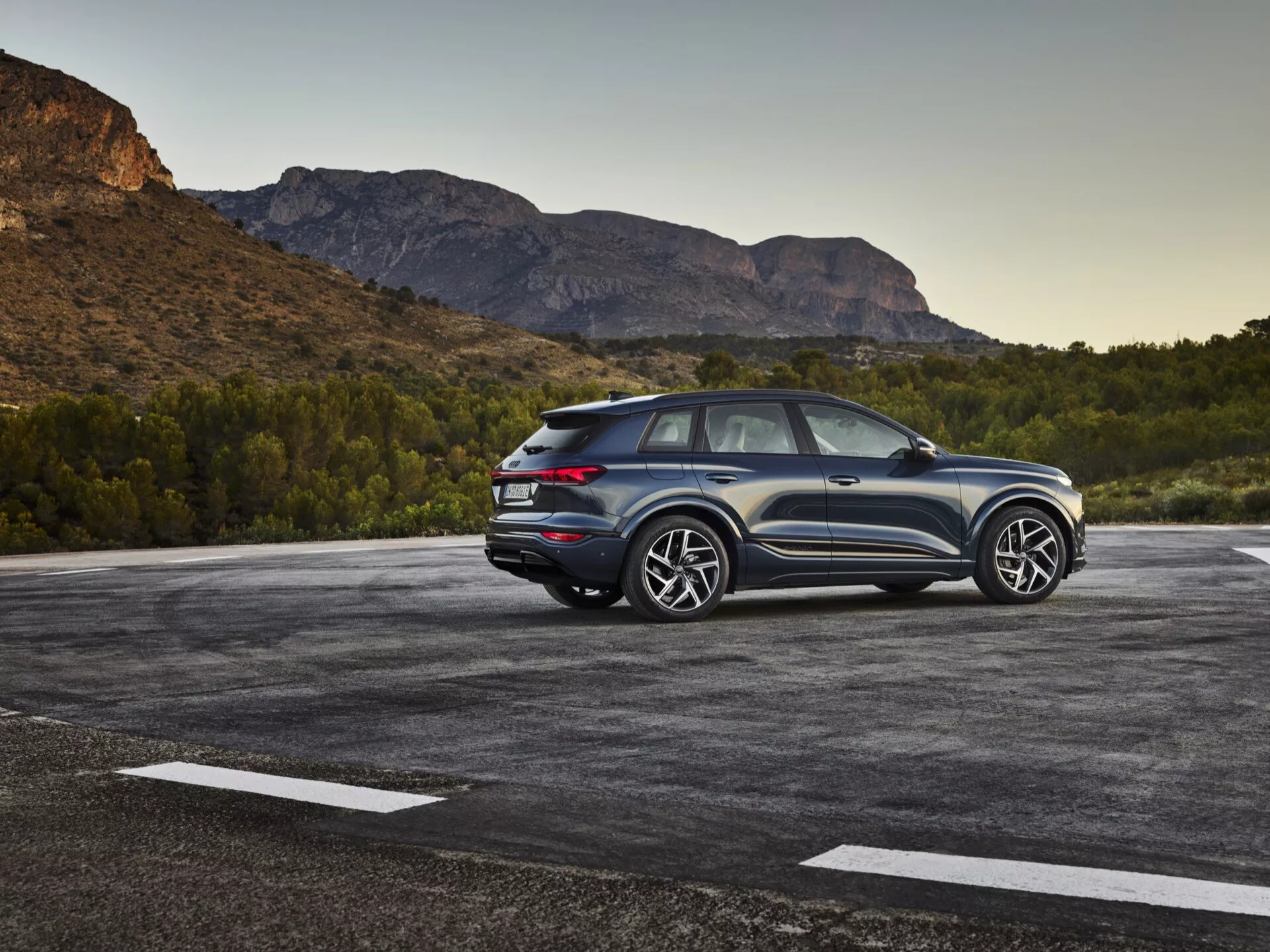 Obraz SUV Audi Q6-e-tron 2024 zaparkowanego na zewnątrz, w tle widać naturalny krajobraz, być może górzysty. Zdjęcie przedstawia widok z trzech czwartych tyłu, podkreślając zarówno boczne, jak i tylne elementy designu. Widoczna jest strona kierowcy, pokazująca linie projektowe, tylne okna i design kół. Pochylony dach sugeruje sportowy, coupe-like design SUV-a. Nowoczesne światła tylne LED, widoczne czujniki lub reflektory na tylnym zderzaku oraz dwukolorowa kolorystyka podkreślają wysokość i postawę. Duże felgi aluminiowe i znaczne opony wskazują na wyższą wersję wyposażenia z sportową estetyką. Samochód wydaje się być blisko kamery, zapewniając szczegółowy widok. Warto zauważyć takie cechy jak oświetlenie LED, czujniki parkowania, koła i opony o wysokiej wydajności, aerodynamiczny design oraz możliwe zaawansowane systemy wspomagania kierowcy i napęd na wszystkie koła.