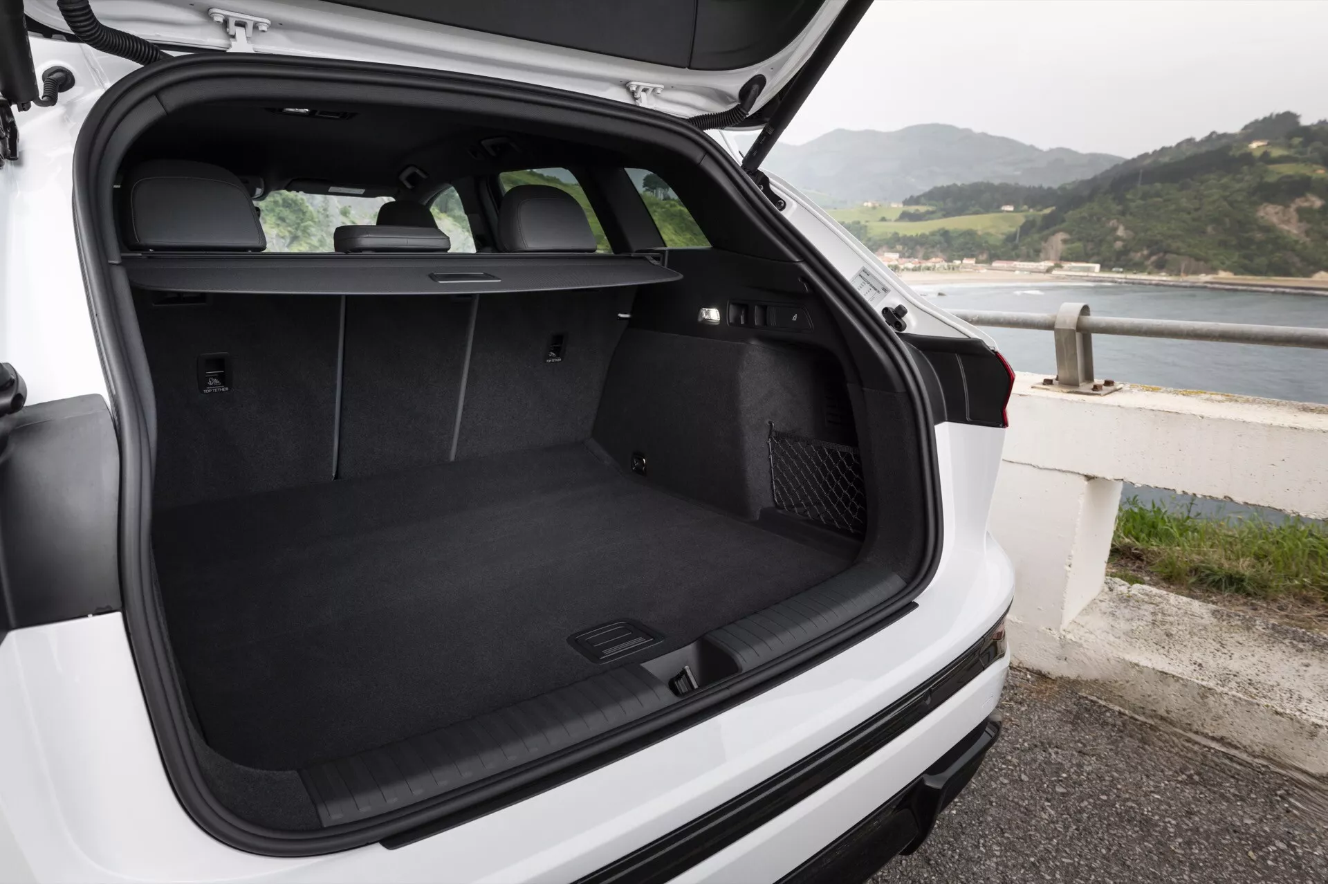 Zdjęcie przedstawia przestrzeń bagażową białego Audi Q6 e-Tron 2024, hatchbacka lub SUV, widzianą od tyłu z otwartą klapą bagażnika i delikatnym nachyleniem kamery w dół. Czarno wykładany bagażnik jest pusty, co pokazuje jego dużą przestrzeń i nienaganny stan. Tylne siedzenia są ustawione pionowo, co wskazuje na możliwość przewożenia pasażerów lub złożenia siedzeń dla większej przestrzeni ładunkowej. Zasłona bagażnika znajduje się zaraz za tylnymi siedzeniami, ale nie jest rozłożona, co umożliwia pełny widok wnętrza bagażnika. Po lewej stronie przestrzeni bagażowej znajduje się gniazdko 12-voltowe, a po prawej stronie wydaje się być schowek prawdopodobnie przeznaczony do przechowywania drobnych przedmiotów. Widoczna jest tylna wycieraczka. Pojazd jest zaparkowany w pobliżu linii brzegowej, co sugeruje lokalizację nadmorską. W bagażniku nie ma żadnych przedmiotów ani ludzi.