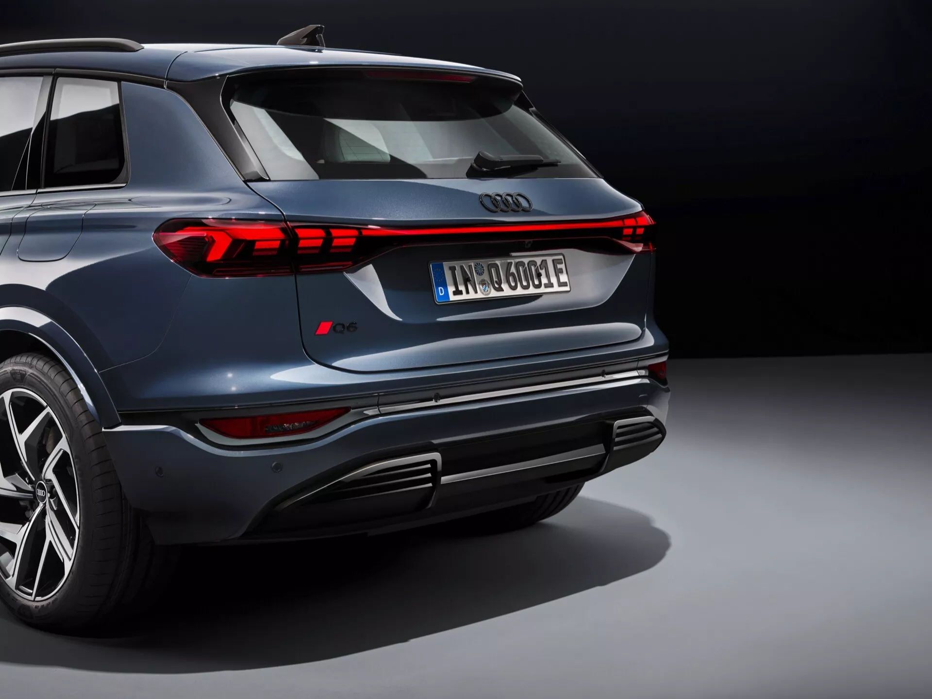 Tylny kąt SUV Audi Q6 e-tron 2024 z metalicznym niebieskim lakierem i nowoczesnymi tylnymi światłami LED. Obraz uwydatnia elegancki design pojazdu ujęciem z niskiego kąta, podkreślając tylny zderzak, zintegrowane końcówki wydechu, dyfuzor i duże felgi aluminiowe. Charakterystyczna pozioma sygnatura świetlna i wyraźny spojler dachowy podkreślają jego sportową estetykę. Europejska tablica rejestracyjna sugerująca elektryczny lub hybrydowy układ napędowy, z widocznym bieżnikiem opon wskazującym na opony sportowe. Uchwycone w ustawieniu studyjnym z kontrolowanym oświetleniem.