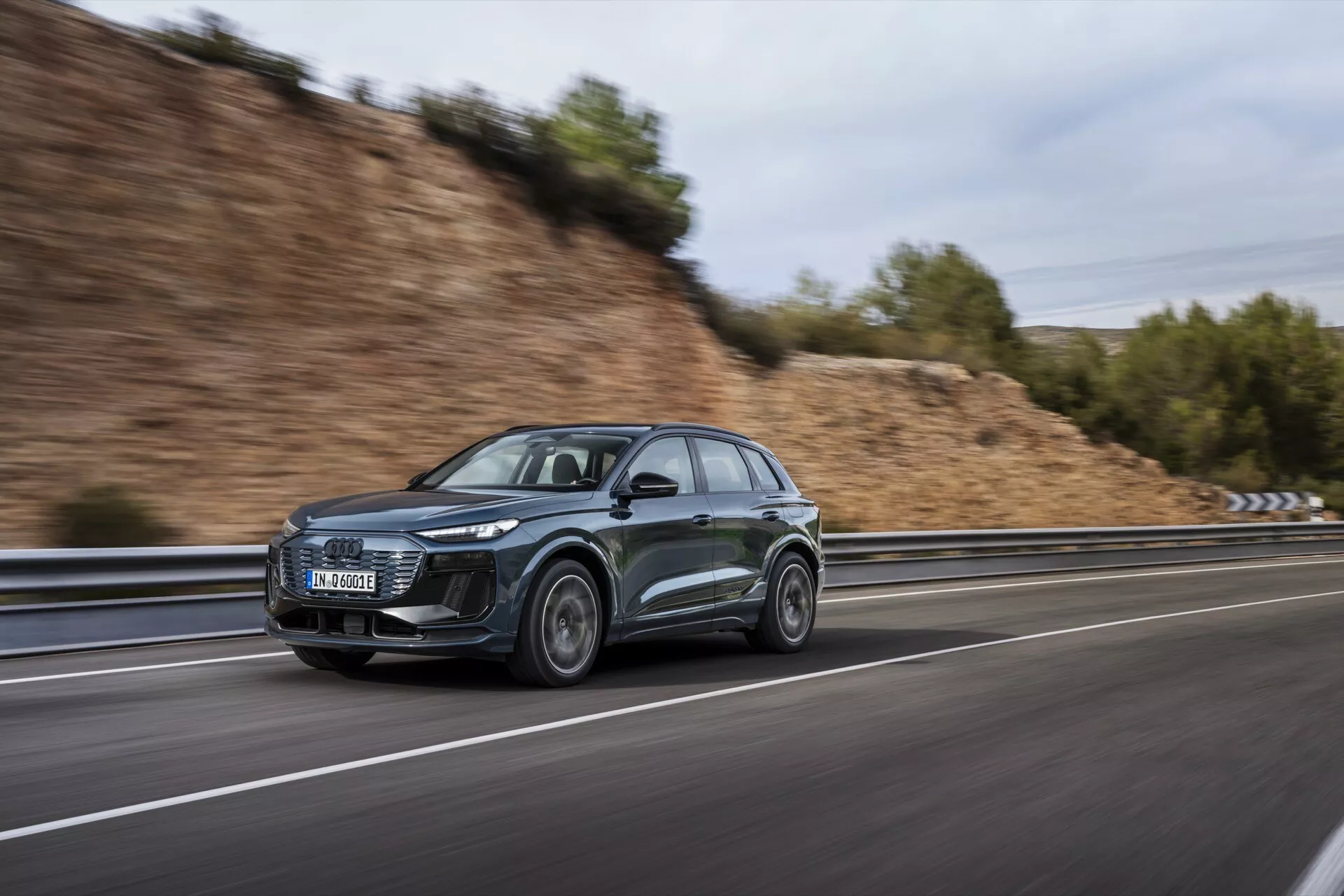 Obraz przedstawia elegancki i nowoczesny SUV Audi Q6 e-tron 2024, sfotografowany z boku podczas ruchu. Nowoczesny design pojazdu podkreślają wyraźne reflektory LED i aerodynamiczne cechy, w tym opadająca linia dachu i opływowe lusterka boczne. Samochód, w ciemnym kolorze, prawdopodobnie szary lub czarny, porusza się po drodze szybkiego ruchu, co widać po rozmytym tle stworzonej za pomocą techniki panoramicznej fotografii. Przednie koło jest lekko skręcone w stronę kamery, ukazując wzór felgi ze stopu. Dodatkowe widoczne elementy obejmują możliwą zintegrowaną technologię w reflektorach i lusterkach bocznych, a także potencjalnie wykorzystanie relingów dachowych.