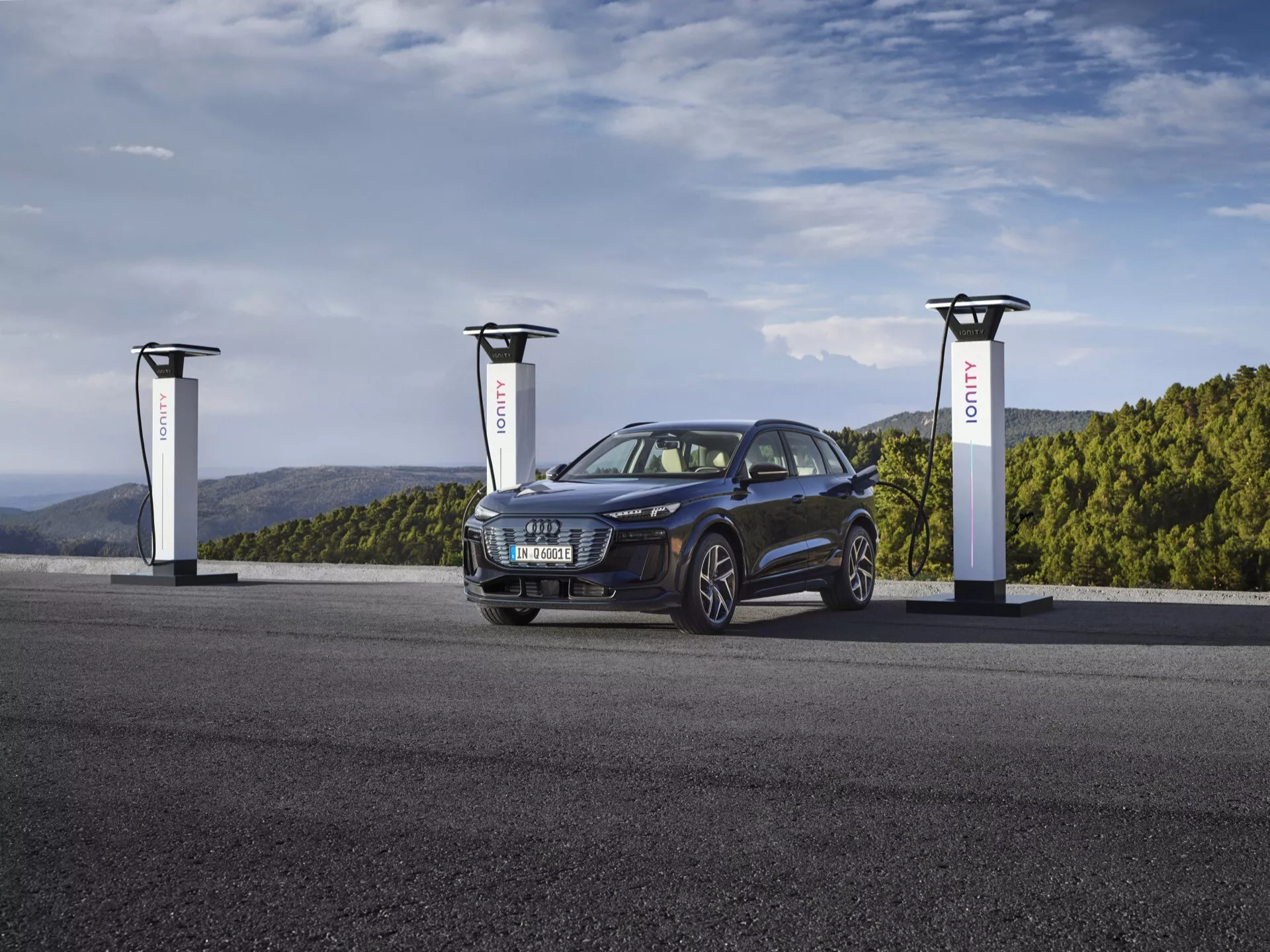 SUV Audi Q6 e-tron 2024 w głębokim metalicznym niebieskim kolorze przy stacji ładowania pojazdów elektrycznych z trzema ładowarkami. Obraz ukazuje pojazd pod kątem trzech czwartych, pokazując przód z lewej strony, podkreślając kratkę, reflektory, koła i maskę. Stylowy i nowoczesny pojazd elektryczny ma sportowy i wszechstronny design z eleganckimi, agresywnymi reflektorami i felgami ze stopów, podkreślając jego zaawansowaną, zrównoważoną technologię motoryzacyjną.