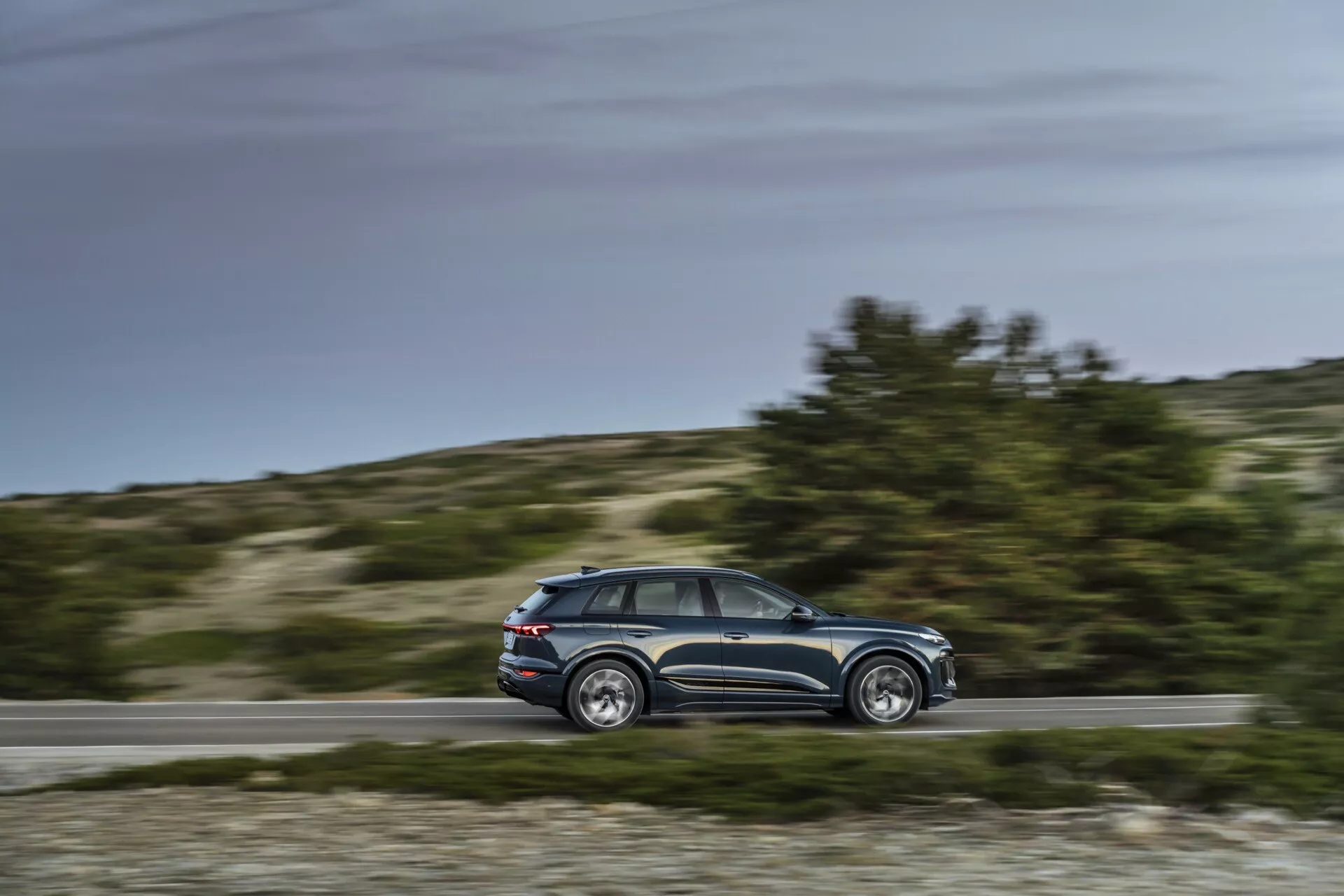 Dynamiczny widok boczny Audi Q6 e-tron 2024 w ruchu z rozmazanym tłem, środkowy kąt równoległy ukazujący profil, linie projektowe, okna, pięć drzwi, lusterka boczne, felgi aluminiowe, ustawienie dzienne, wiejskie tło.