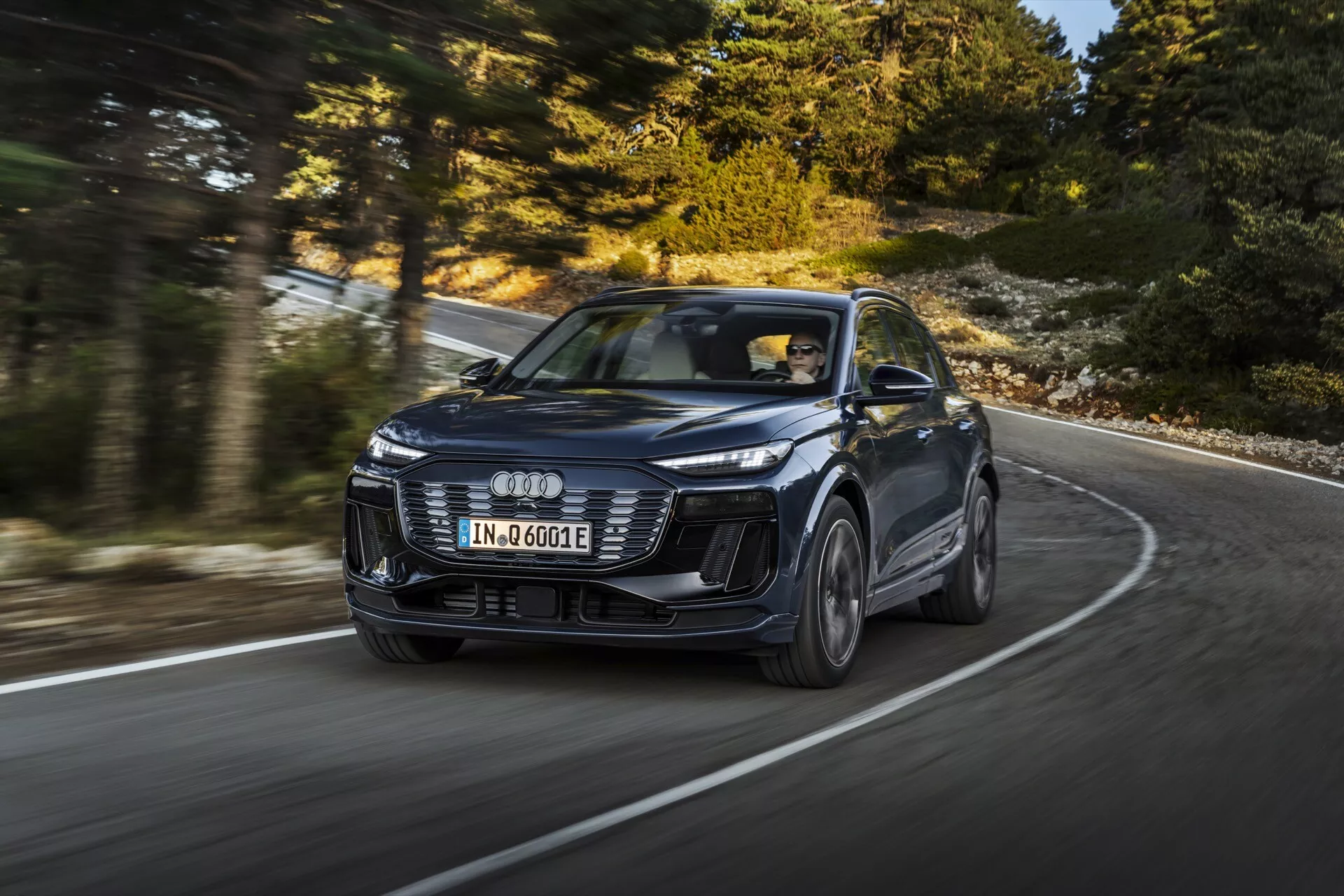 Obraz nowoczesnego SUV-a Audi Q6 e-tron 2024 w ruchu, uchwycony pod dynamicznym kątem trzy czwarte przodu, podkreślający jego design i osiągi. Pojazd, rozpoznawalny po charakterystycznym logo z czterema pierścieniami, posiada emblematyczne reflektory LED, agresywny przedni zderzak i rzeźbione linie maski. Obraz, zrobiony pod niższym kątem, podkreśla obecność auta na zakręconej drodze, z lekko rozmytymi kołami ze stopu wskazującymi na ruch. Nowy model sugeruje zaawansowane technologie, takie jak adaptacyjny tempomat i napęd elektryczny, oznaczone odznaką 'e-tron'. Profesjonalna sesja fotograficzna pokazująca luksus, design i technologie w konfiguracji z kierownicą po lewej stronie.