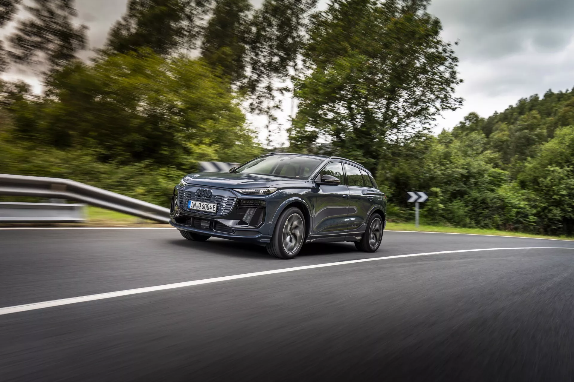 Na zdjęciu widać nowoczesny SUV Audi Q6 e-tron 2024 w ruchu, uchwycony pod kątem od przedniej lewej strony, koncentrując się na lewym boku. Dynamiczne ujęcie podkreśla widoczny przedni lewy reflektor z charakterystycznym dziennym oświetleniem LED, dużą współczesną osłonę chłodnicy, eleganckie aerodynamiczne lusterka boczne z wbudowanymi kierunkowskazami oraz duże felgi ze stopów lekkich z oponami niskoprofilowymi. Samochód posiada panoramiczny dach i przyciemniane szyby, a metaliczny lakier odbija otoczenie. Niski punkt widzenia i lekkie rozmycie tła podkreślają szybkość i ruch samochodu na zakrzywionej dwupasmowej drodze.