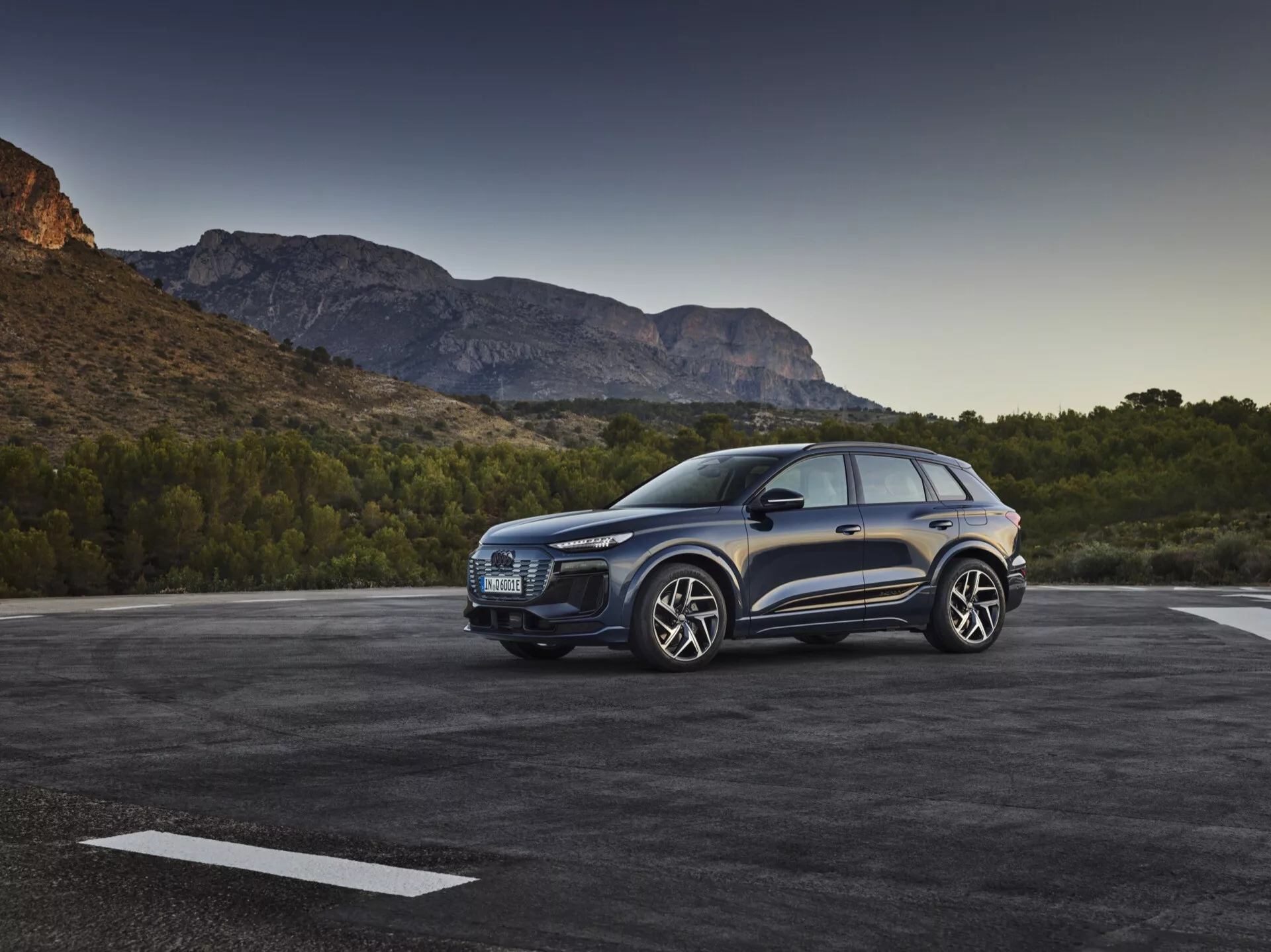 Trzy czwarte widok z przodu niebieskiego SUV-a Audi Q6 e-tron 2024 zaparkowanego na zewnątrz, ukazującego charakterystyczny grill, eleganckie reflektory, rzeźbioną maskę, duże felgi aluminiowe, lusterka boczne i drzwi po stronie kierowcy, na tle malowniczej górskiej scenerii przy porannym lub późno popołudniowym świetle.
