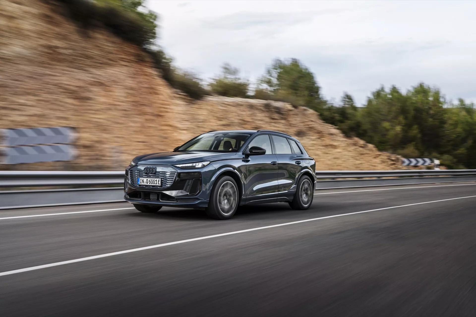 Obraz Audi Q6 e-tron 2024 w ruchu, uchwycony za pomocą techniki panningu, która podkreśla elegancki profil i nowoczesny design SUV-a. Samochód, sfotografowany z boku, pędzi po wielopasmowej autostradzie z rozmytymi kołami i tłem, co podkreśla jego dynamiczne możliwości. Ciemne metaliczne auto ma dużą przednią kratkę, charakterystyczne reflektory LED, sportowe felgi aluminiowe i odblaskowe powierzchnie. Zachmurzone niebo i mokra droga tworzą dramatyczną scenę, podkreślając luksusowe i wydajne cechy pojazdu.