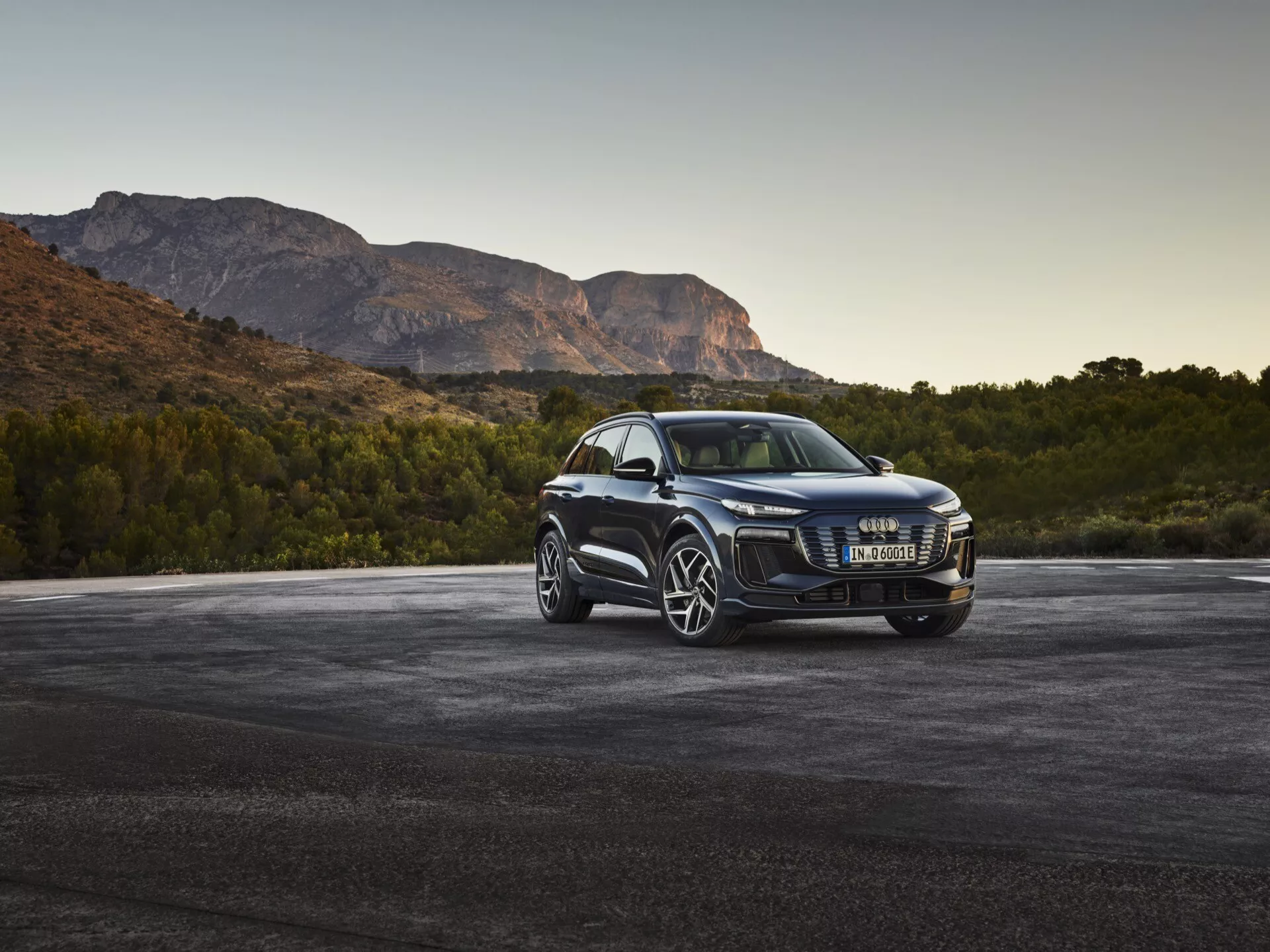 Luksusowy SUV Audi Q6 e-tron 2024 w trójćwierciowym froncie, z widoczną chromowaną kratką, nowoczesnymi reflektorami LED, dużymi wieloramiennymi felgami ze stopów metalowych, bocznymi lusterkami z wbudowanymi kierunkowskazami, przyciemnianymi szybami i lśniącym wykończeniem, zaparkowany na asfalcie z górami w tle o świcie lub zmierzchu.