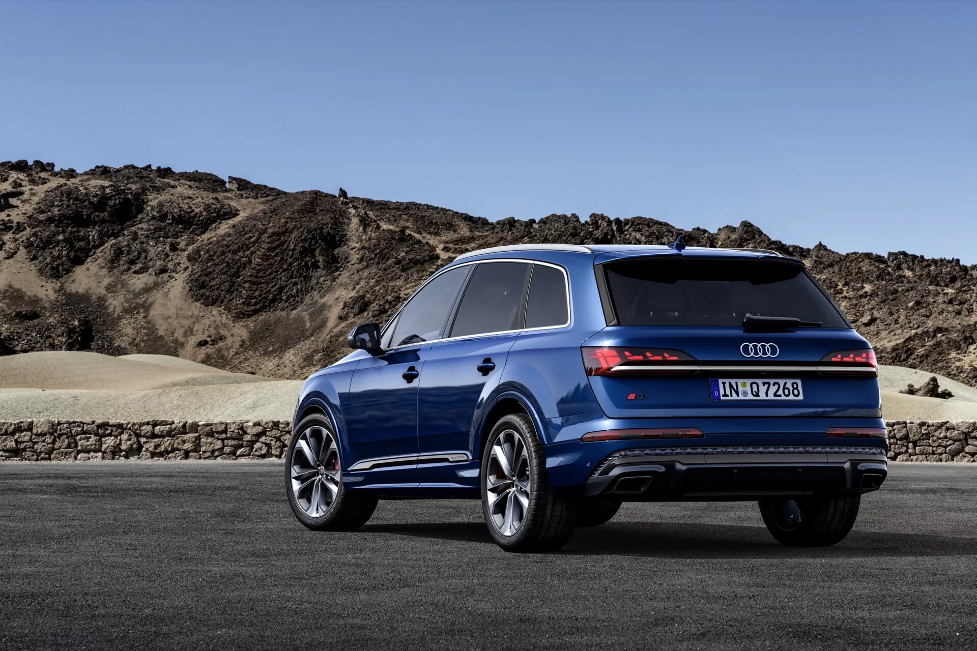 Boczny tylny widok Audi Q7 2024 (4M-Facelift-2) pokazujący błyszczącą niebieską powłokę, nowoczesny design tylnych świateł sięgających do klapy bagażnika, podwójny wydech i tylny dyfuzor dla lepszej aerodynamiki, duże felgi z wieloramienne wzorem i niskoprofilowymi oponami dla wysokiej wydajności na drodze, panoramiczny dach, relingi dachowe na dodatkowy bagaż i przyciemniane tylne okno, na tle półpustynnego krajobrazu.