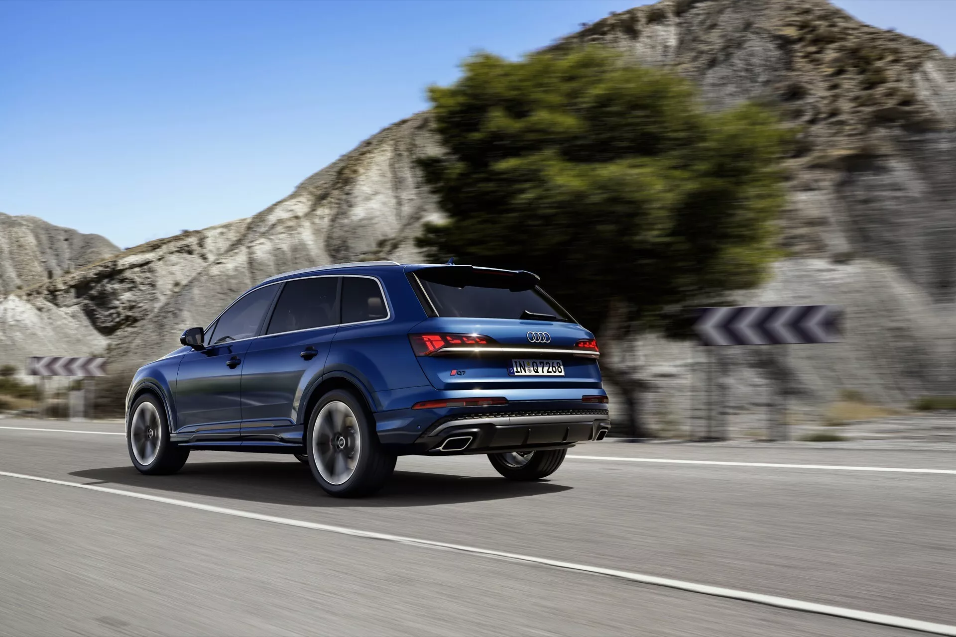 Dynamiczny widok z tyłu ukośnie na niebieskiego luksusowego SUV-a Audi Q7 z 2024 roku w ruchu na krętej drodze z tłem skalistego zbocza, podkreślający linie wzornicze, sportowe tylne światła, podwójny wydech i zaawansowane funkcje technologiczne