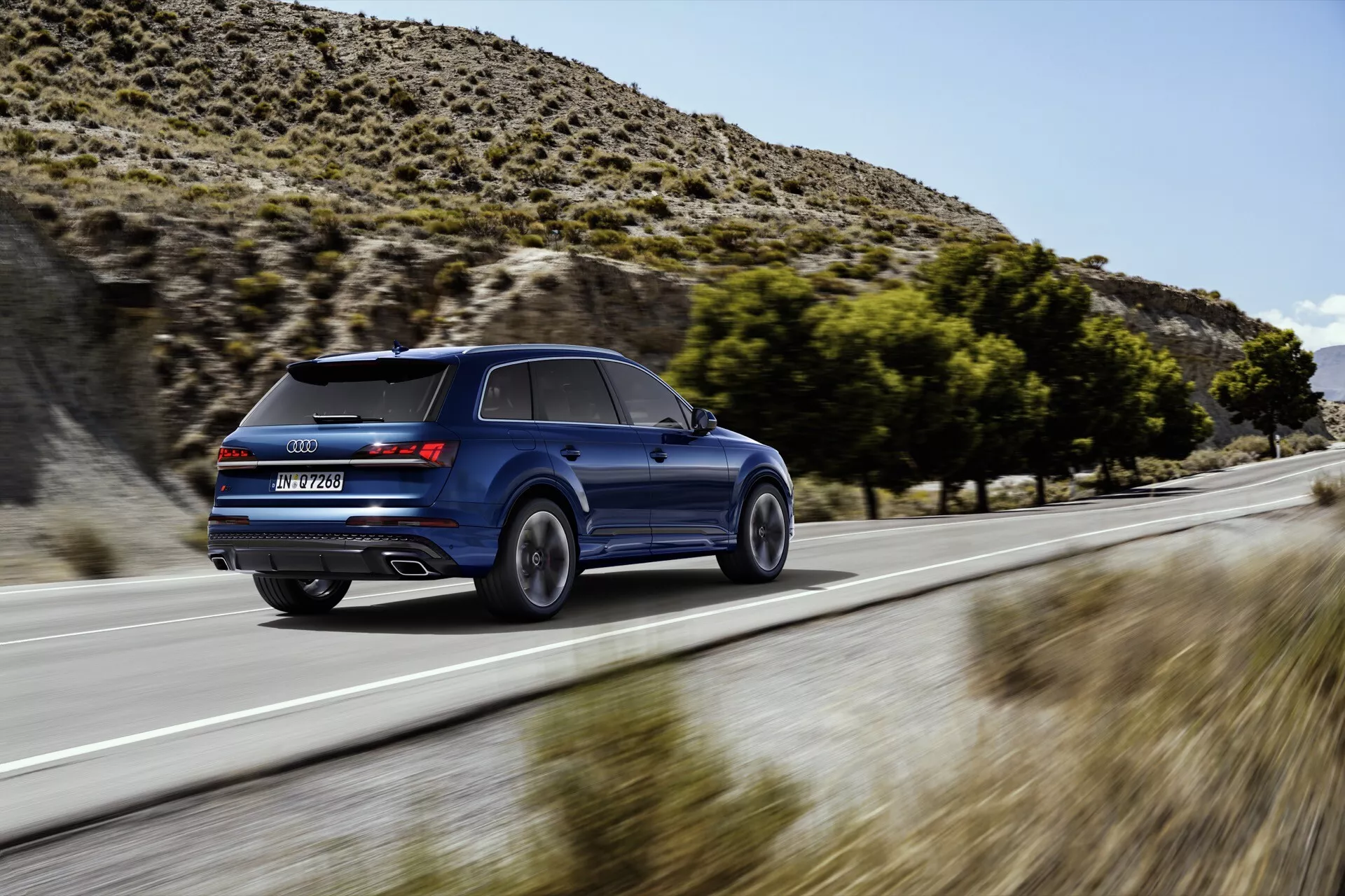 Audi Q7 2024 (4M Facelift 2) w ruchu, pokazane z dynamicznej perspektywy trzy czwarte z tyłu, podkreślające jego wysokość, elegancki design, poszerzone nadkola i nachylony tył dachu. Metalicznie niebieskie SUV podkreśla swoje osiągi rozmyciem w ruchu, tylnymi światłami LED, spojlerem dachowym, podwójnymi portami wydechowymi i unikalnym wzorem kół, sugerując zaawansowaną technologię i luksus.