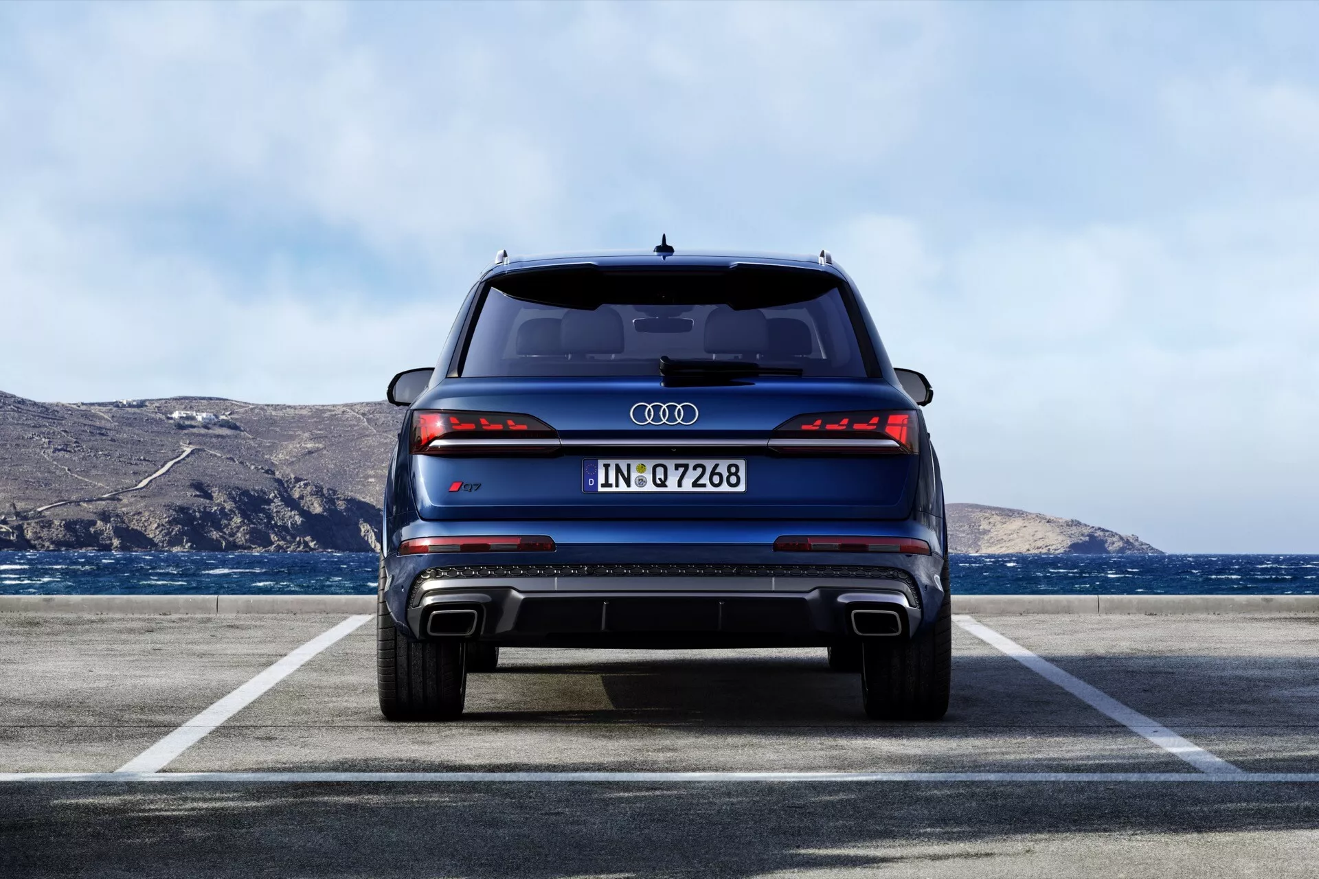Tylny widok nowoczesnego SUV-a Audi Q7 2024 (4M-Facelift-2) zaparkowanego na zewnętrznym parkingu z krajobrazem wybrzeża w tle, prezentujący tylne światła LED, podwójny wydech, dyfuzor tylny, czujniki parkowania, wysoko umieszczone światło stop, spoiler dachowy i potencjalne funkcje kamery cofania w jasnych warunkach dziennych