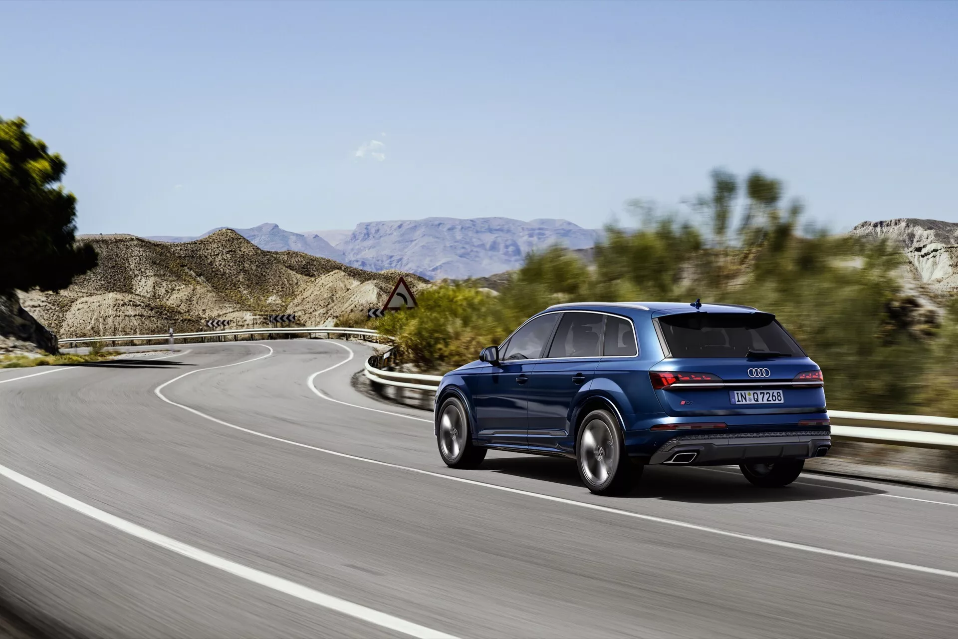 Audi Q7 SUV 2024 w ruchu na krętej drodze, pokazany w ciemnoniebieskim kolorze metalicznym, widok z tyłu pod kątem trzech czwartych z widocznymi światłami tylnymi, zderzakiem tylnym, podwójnymi końcówkami wydechu i premium felgami aluminiowymi pod jasnym niebem z dalekimi górami.