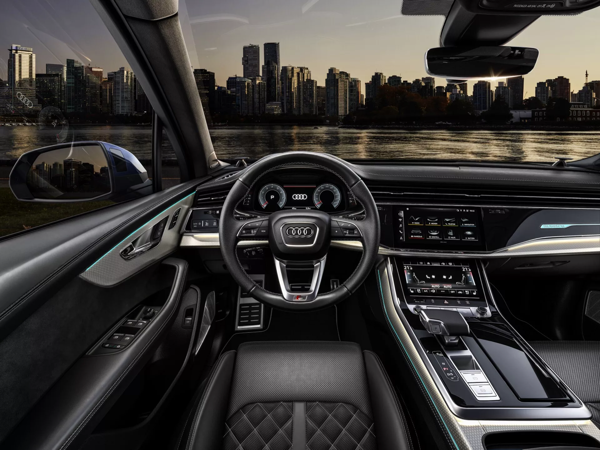 Widok wnętrza Audi Q7 2024 (4M-Facelift-2) z miejsca kierowcy pokazujący wielofunkcyjną kierownicę pokrytą skórą ze znakiem, cyfrowy zestaw wskaźników, duży poziomy system informacyjno-rozrywkowy z ekranem dotykowym, elegancki design wnętrza z materiałów premium, sterowanie dotykowe na konsoli środkowej, pikowane fotele premium i oświetlenie ambientowe, wraz z widokiem na linię horyzontu miasta przez przednią szybę