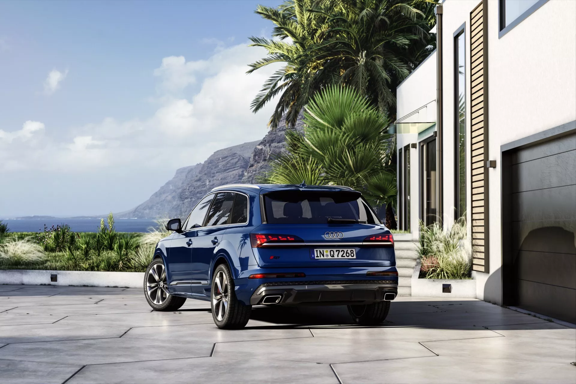 Luksusowy SUV Audi Q7 2024 (4M Facelift 2) w metalicznym niebieskim kolorze, zaparkowany na przydomowym podjeździe z widokiem na ocean, z sportowym wyglądem tylnej części, nowoczesnymi światłami LED, luksusowymi alufelgami i podwójnymi końcówkami wydechu.