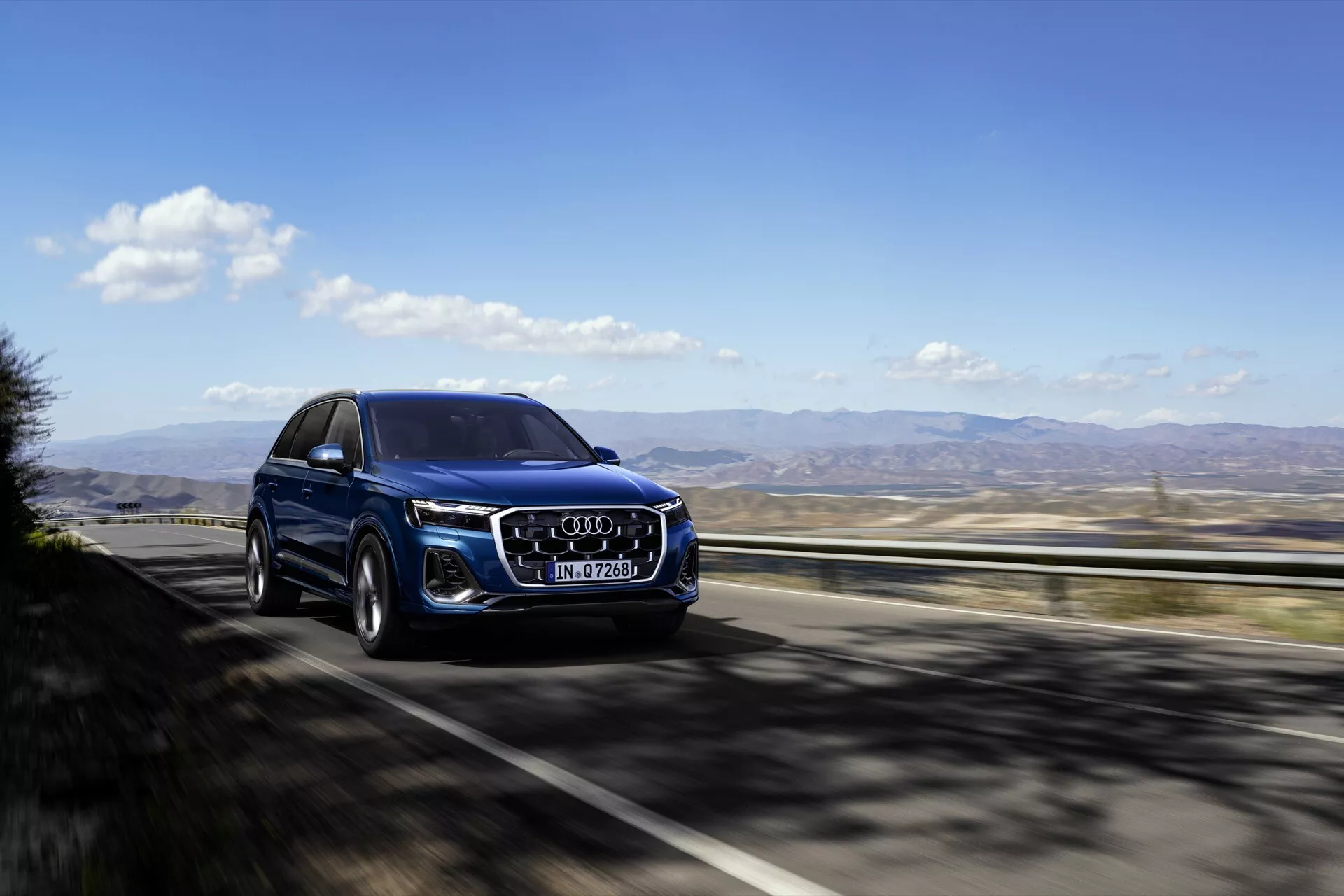 Audi Q7 2024 (4M-Facelift-2) w metalicznym niebieskim kolorze, podróżujące autostradą, prezentuje swój przód pod kątem trzech czwartych z charakterystyczną atrapą Audi, reflektorami LED, dużymi felgami ze stopów i relingami dachowymi na tle jasnego nieba i górzystego terenu.