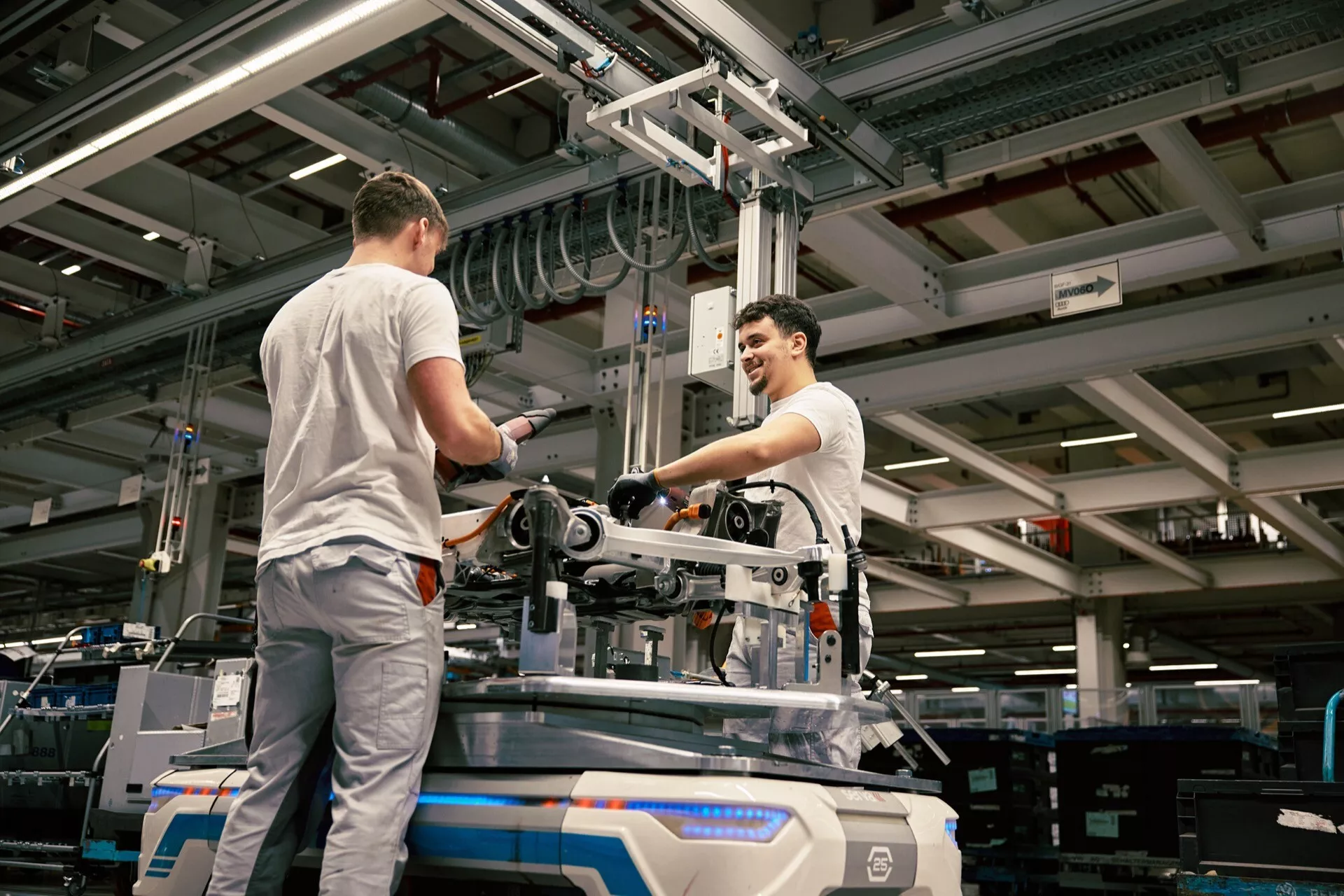 Sceneria przemysłowa z dwoma pracownikami montującymi lub konserwującymi sprzęt mechaniczny na podłodze fabryki, pod sztucznym oświetleniem, z podwyższonym punktem widzenia podkreślającym środowisko produkcyjne