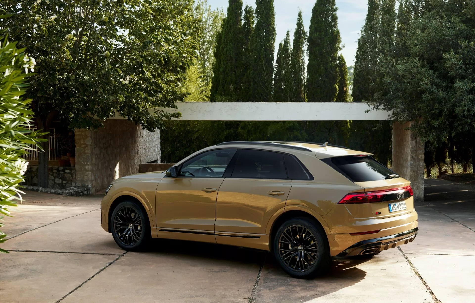 Luksusowy SUV Audi Q8 (4M-Facelift) z 2023 roku zaparkowany na ekskluzywnym podjeździe, prezentujący sportowy tylny spoiler, elegancką linę dachu i nowoczesne tylne światła LED, w metalicznym złotym wykończeniu z odbiciami na błyszczącej karoserii, otoczony drzewami i luksusową strukturą w tle