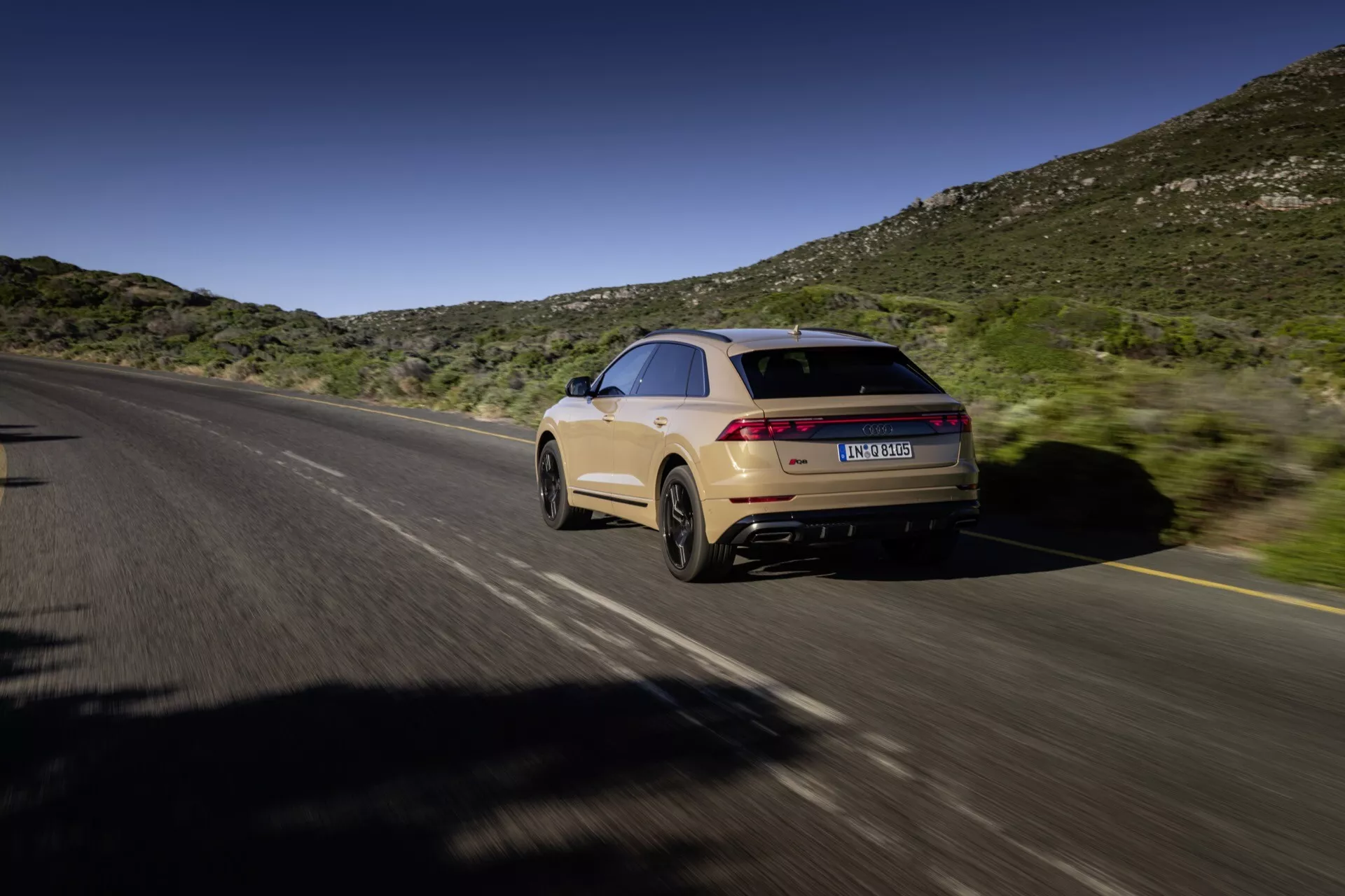 Audi Q8 2023 (4M-Facelift) w ruchu, uchwycone z perspektywy tyłu i boku na podzielonej drodze z tłem naturalnego krajobrazu wzgórz pod niebieskim niebem, pokazując linię dachu typu coupé, elegancki design, wydłużone światła tylne, jasnobrązowy lakier i podwójny system wydechowy.