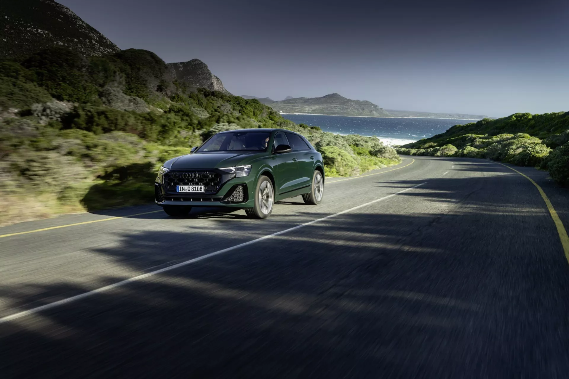 Audi Q8 2023 (4M-Facelift) jadące drogą przybrzeżną, prezentujące widok z przodu i boku na jego dużą kratę, reflektory LED, elegancką maskę i częściową boczną klapę, uosabiające technologię i luksus w ruchu na tle czystego nieba, gór i oceanu.