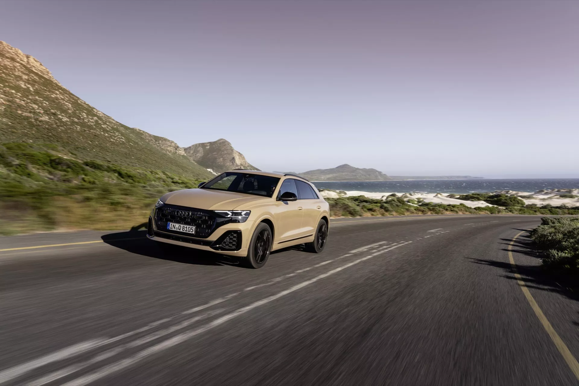 Widok z przodu na trzy czwarte Audi Q8 2023 (4M-Facelift) na nadmorskiej drodze, ukazujący reflektory LED, aerodynamiczny design i rozmycie ruchu wskazujące na ruch