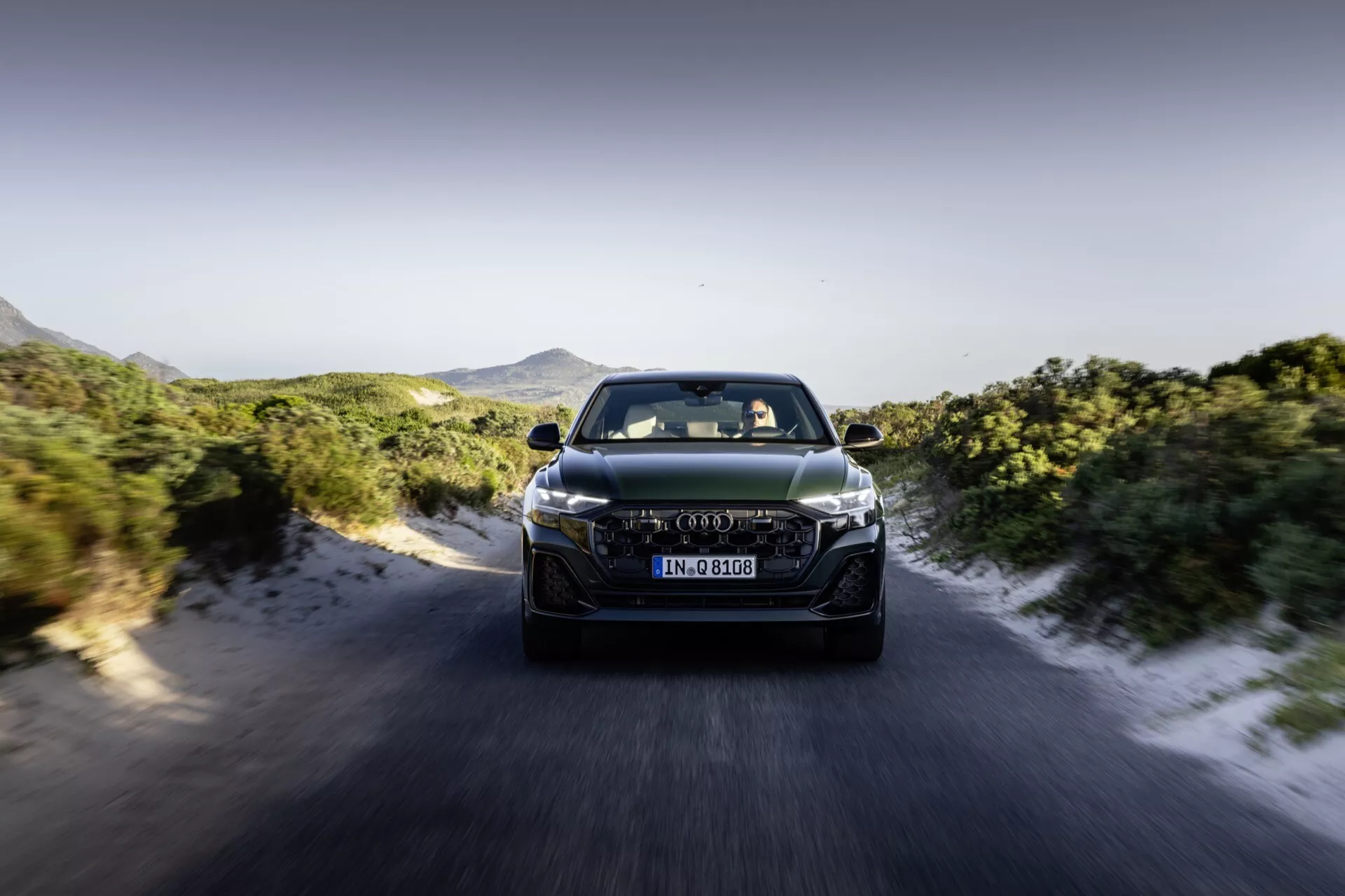 Audi Q8 4M-Facelift 2023 w ruchu, widok z przodu prezentujący reflektory LED, zaawansowany design grilla i cechy aerodynamiczne na tle dynamicznej drogi i górskiego krajobrazu