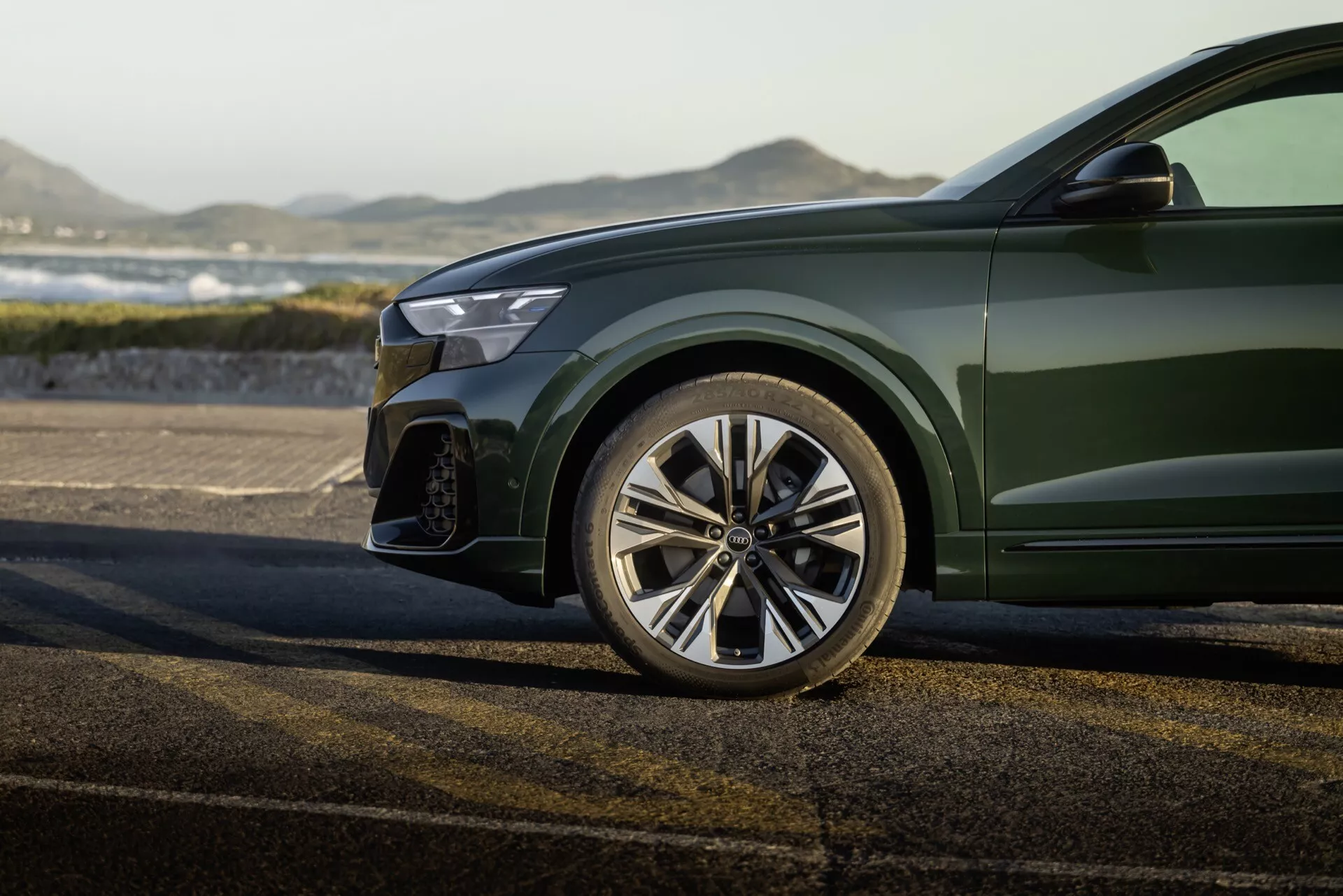Zbliżony widok sportowego przedniego lewego kwartału Audi Q8 2023 (4M-Facelift), pokazujący polerowane wieloramienne koło aluminiowe, częściowy przedni zderzak z dużym wlotem powietrza, krawędź nowoczesnego reflektora LED i błyszczący ciemnozielony metaliczny lakier karoserii, na tle krajobrazu zewnętrznego.