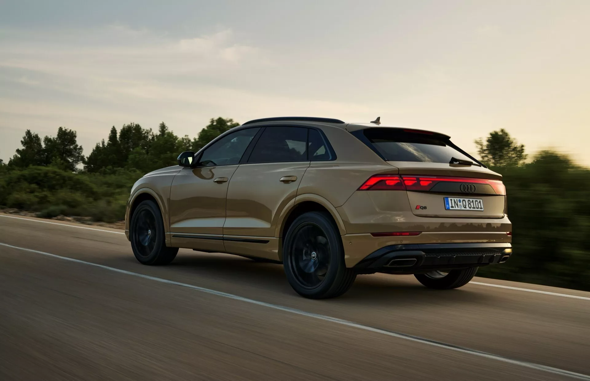 Widok z tyłu trzy czwarte SUV-a Audi Q8 (4M-Facelift) 2023 w ruchu, ukazujący charakterystyczny design świateł tylnych, linie konturu i profil koła, uchwycony podczas dynamicznej jazdy w wiejskim otoczeniu przy ciepłym oświetleniu.