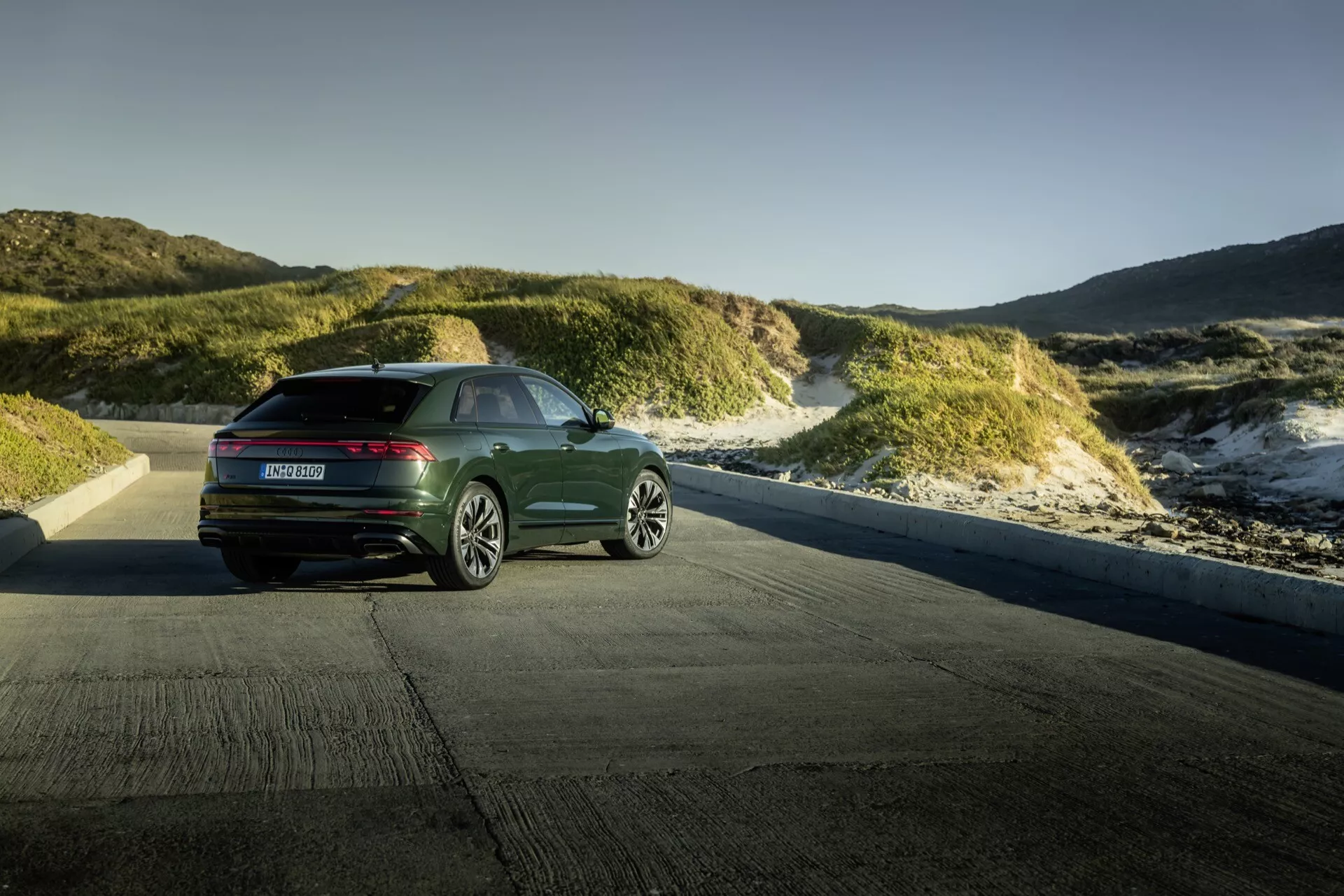 Audi Q8 4M-Facelift 2023 zaprezentowane na tle nadmorskiego krajobrazu wydm podczas złotej godziny, pokazujące widok z tyłu i boku z nowoczesnymi poziomymi światłami tylnymi i eleganckim designem