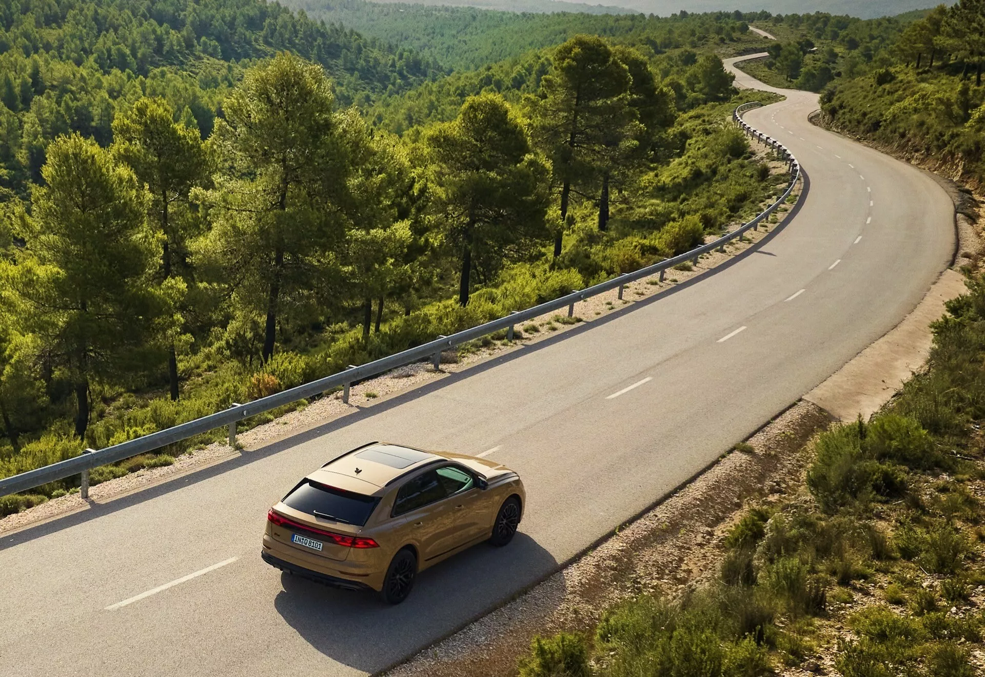 Złote Audi Q8 2023 (4M-Facelift) na krętej drodze dwupasmowej w wiejskim lub leśnym otoczeniu, widziane z podniesionego kąta ukazujące tylną i boczną część jego konstrukcji na tle naturalnego krajobrazu