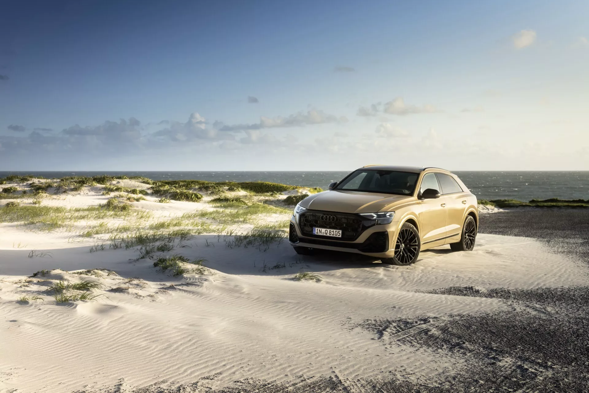Audi Q8 2023 (4M-Facelift) zaparkowane na piaszczystym terenie pod częściowo zachmurzonym niebem z widokiem na morze w tle, prezentujące przedni grill, reflektory LED i aerodynamiczny design