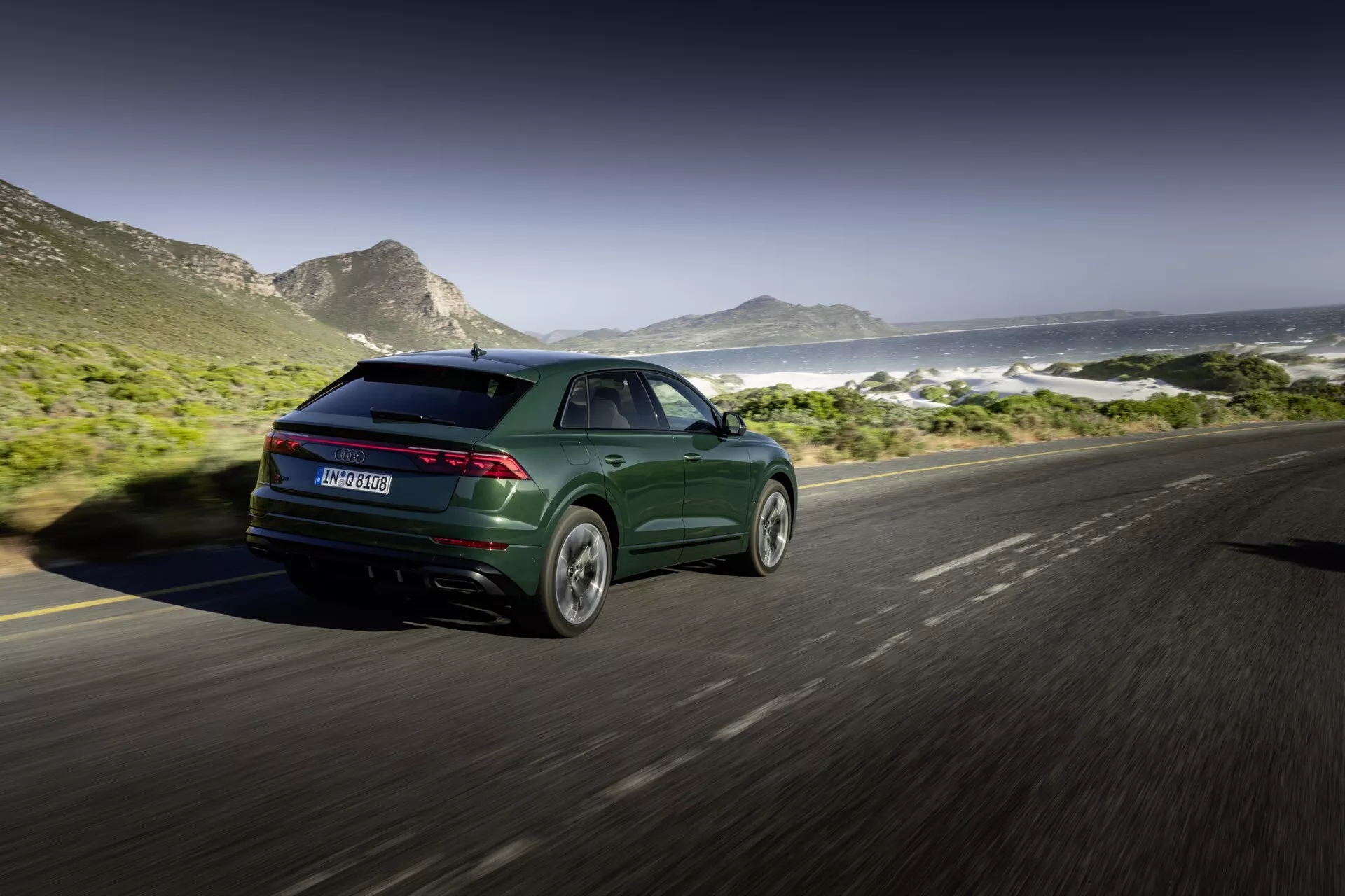 Audi Q8 2023 (4M-Facelift) w metalicznym ciemnozielonym kolorze, prezentujące sportowy i nowoczesny design z eleganckimi światłami tylnymi i alufelgami, uchwycone z tylnego ujęcia trzy-czwarte na nadmorskiej drodze z górami i morzem w tle
