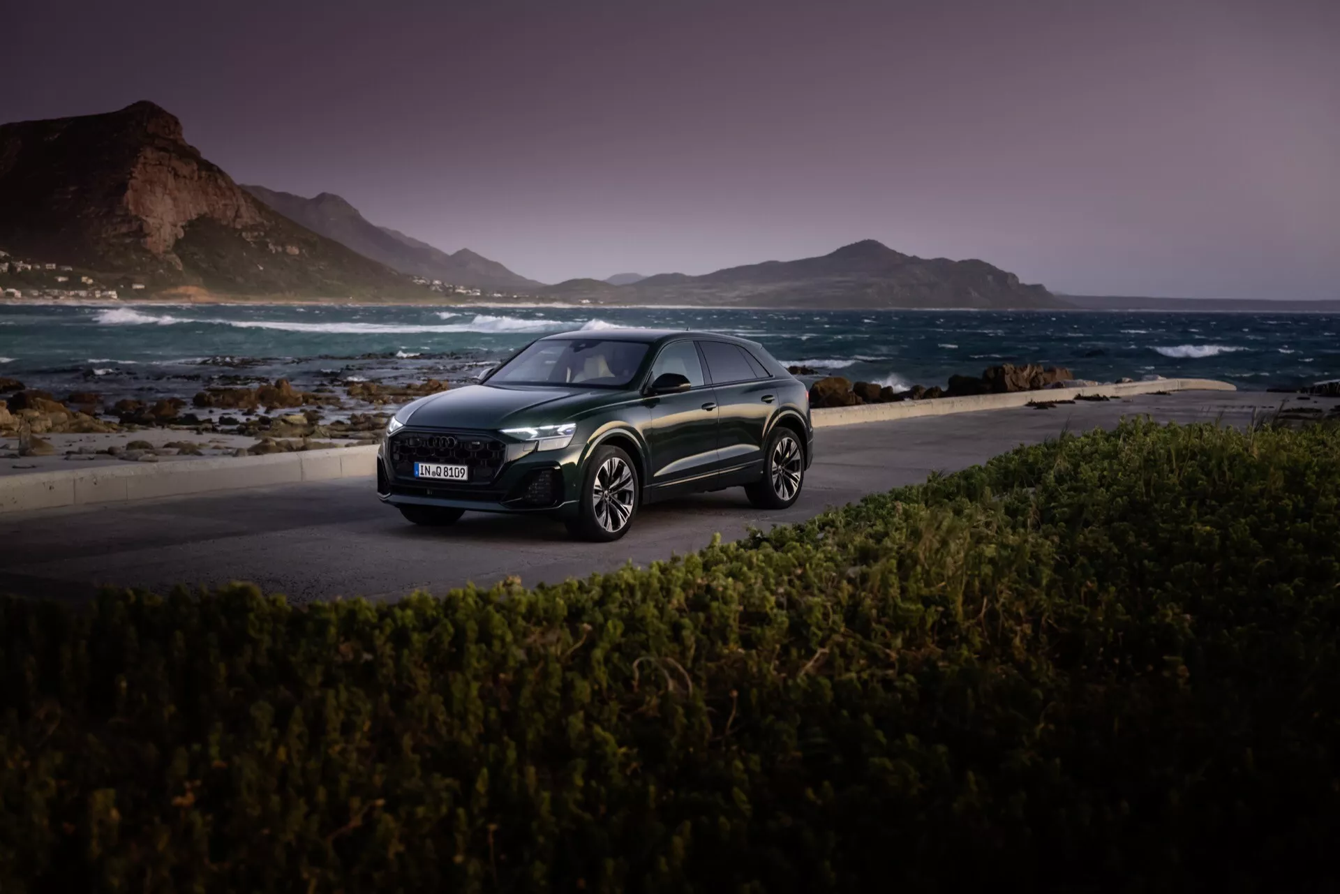 Audi Q8 2023 (4M-Facelift) ciemny hatchback na malowniczej przybrzeżnej drodze o zmierzchu, pokazujący jego reflektory LED, aerodynamiczny profil i obręcze z stopów lekkich