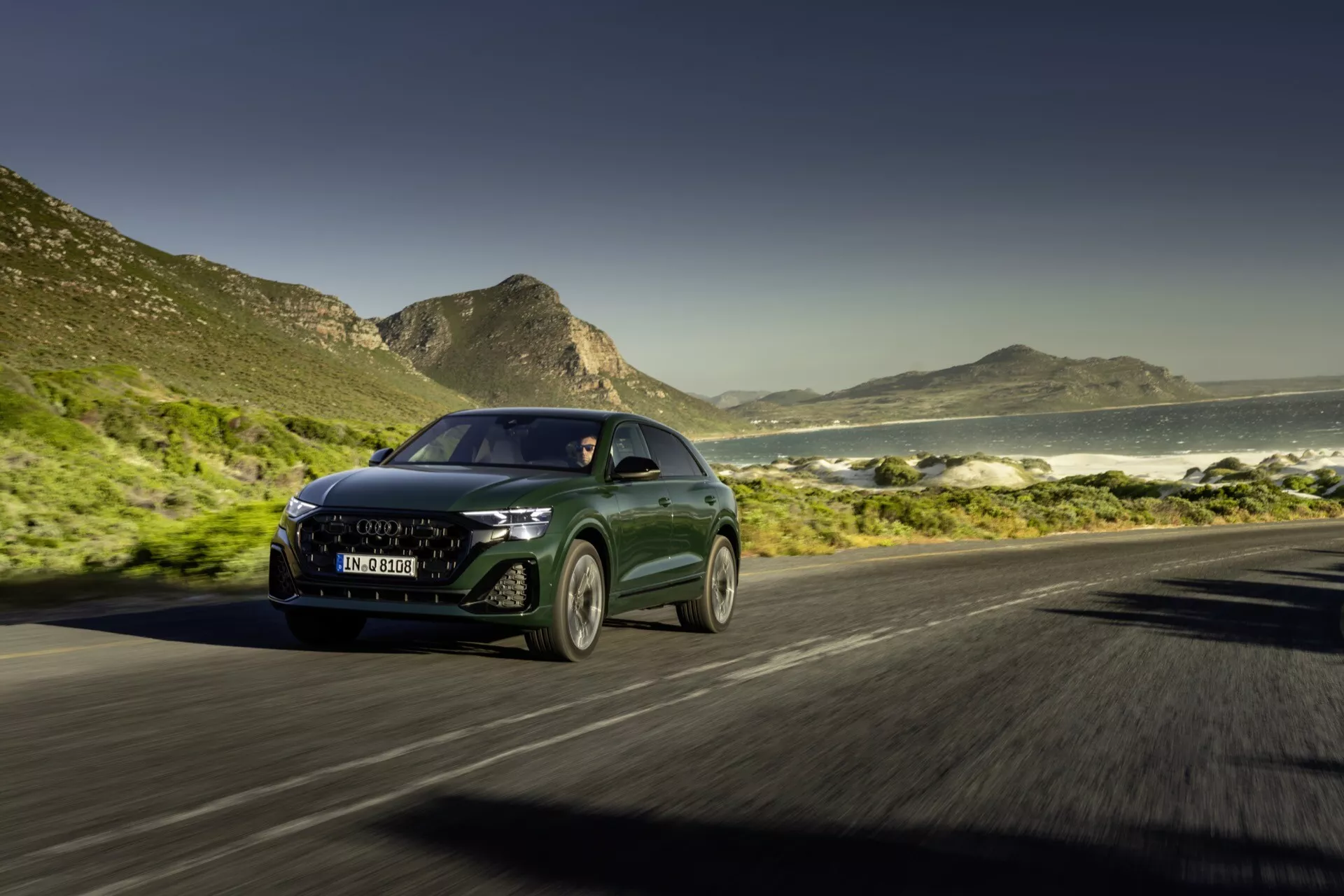 Audi Q8 4M Facelift 2023 jedzie po nadmorskiej drodze, dynamiczny przód z trzech czwartych z widocznymi reflektorami LED, nowoczesną atrapą i efektem rozmycia ruchu na tle oceanu i gór w słoneczny dzień