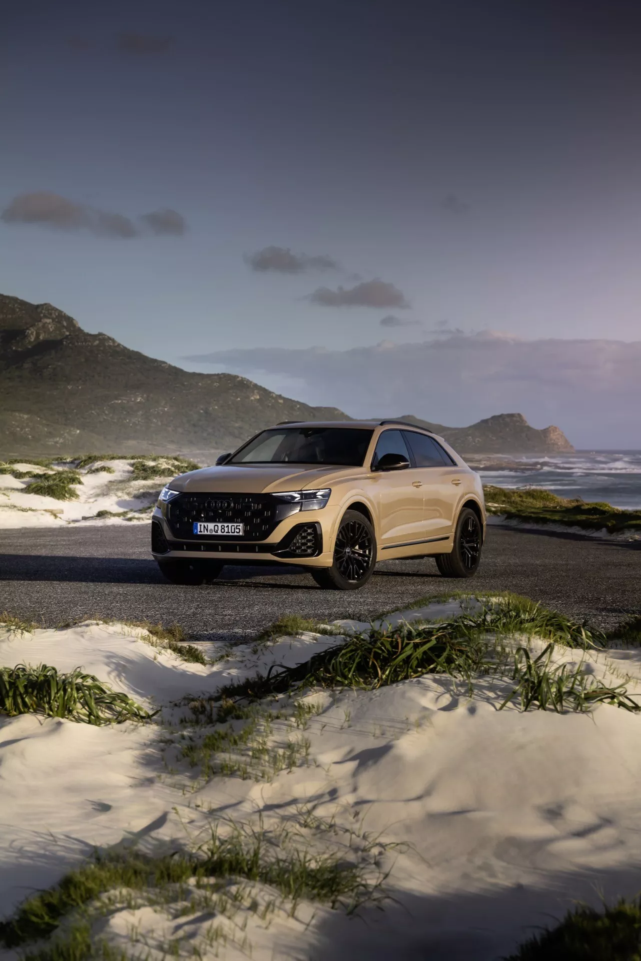 Audi Q8 2023 (4M-Facelift) w majestatycznym krajobrazie plaży i gór o zmierzchu, prezentujące swój elegancki design i zaawansowane funkcje jak LEDowe reflektory i duże felgi aluminiowe, z kąta trzy czwarte z przodu i boku