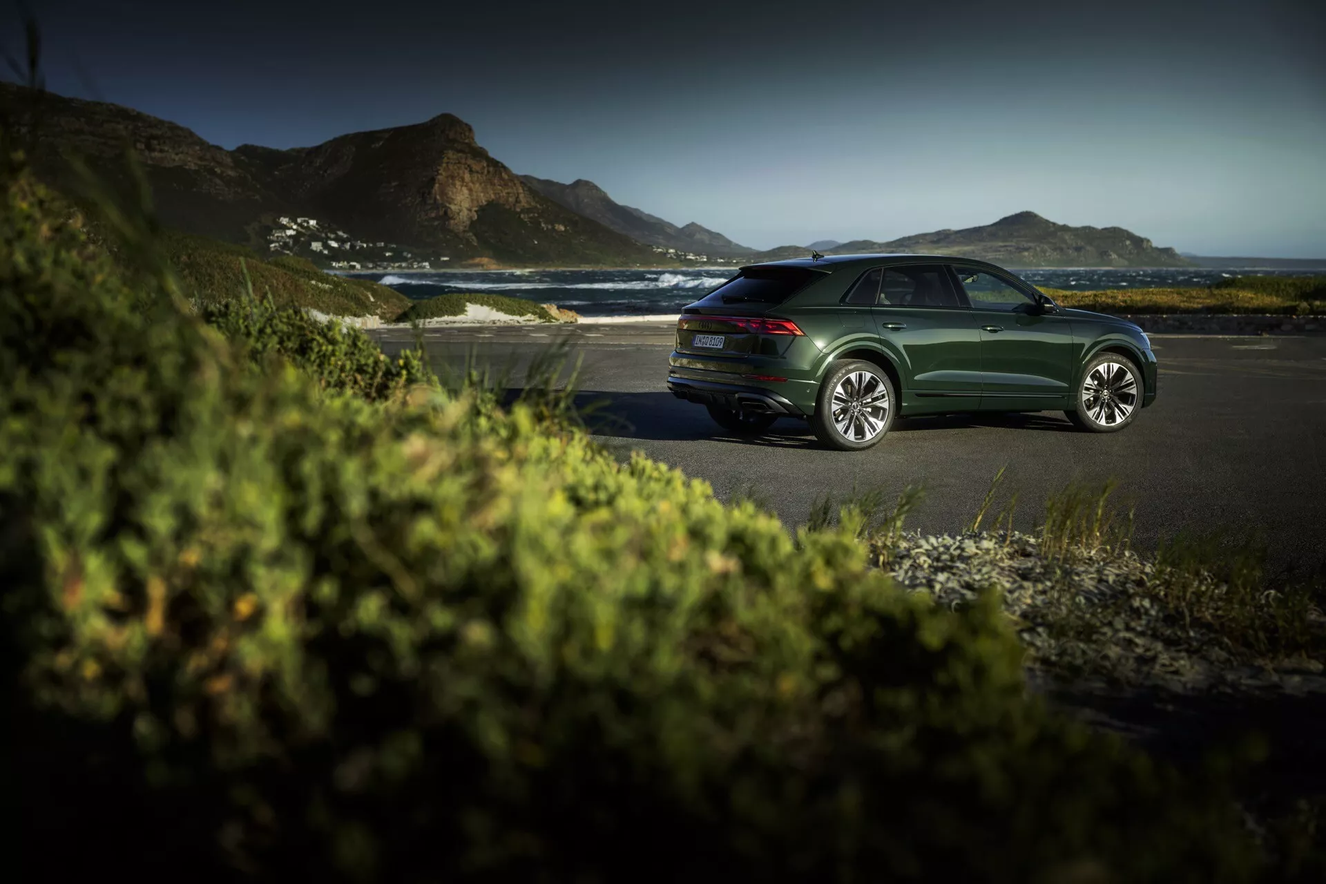 Sedan Audi Q8 (4M-Facelift) w ciemnozielonym kolorze zaparkowany na przybrzeżnej drodze podczas złotej godziny, prezentujący eleganckie linie wzornicze, wieloramienne koła ze stopu i spokojne, luksusowe otoczenie na zewnątrz