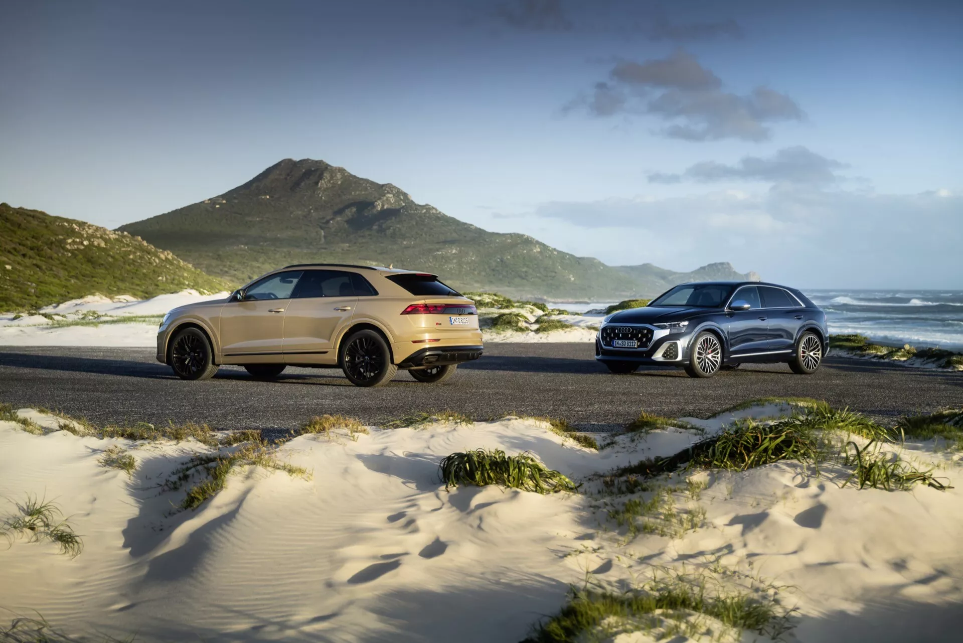 Modele Audi Q8 (4M-facelift) z 2023 roku zaprezentowane na piaszczystej plaży, z złotym sportowym SUV-em po lewej stronie w widoku z trzech czwartych od tyłu i srebrnym tradycyjnym SUV-em po prawej stronie w widoku z trzech czwartych od przodu, na tle wydm trawiastych, gór i częściowo pochmurnego nieba