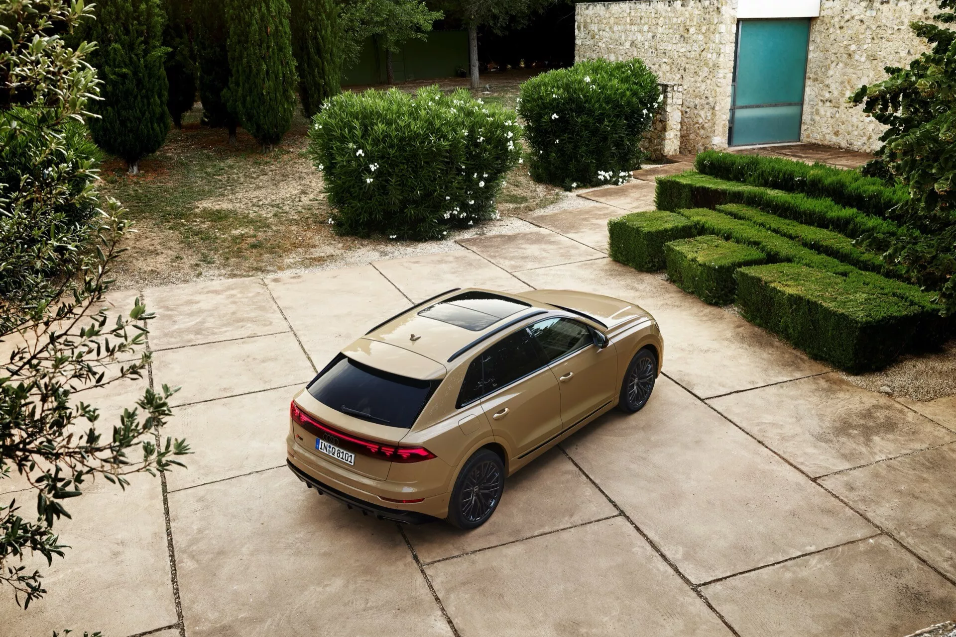 Sedan Audi Q8 2023 (4M-facelift) z wysokiego punktu perspektywy od tyłu i boku, ukazująca LEDowe światła tylne, panoramiczny dach oraz sportowe felgi, zaparkowany z malowniczym tłem żywopłotów i budynku z kamienia