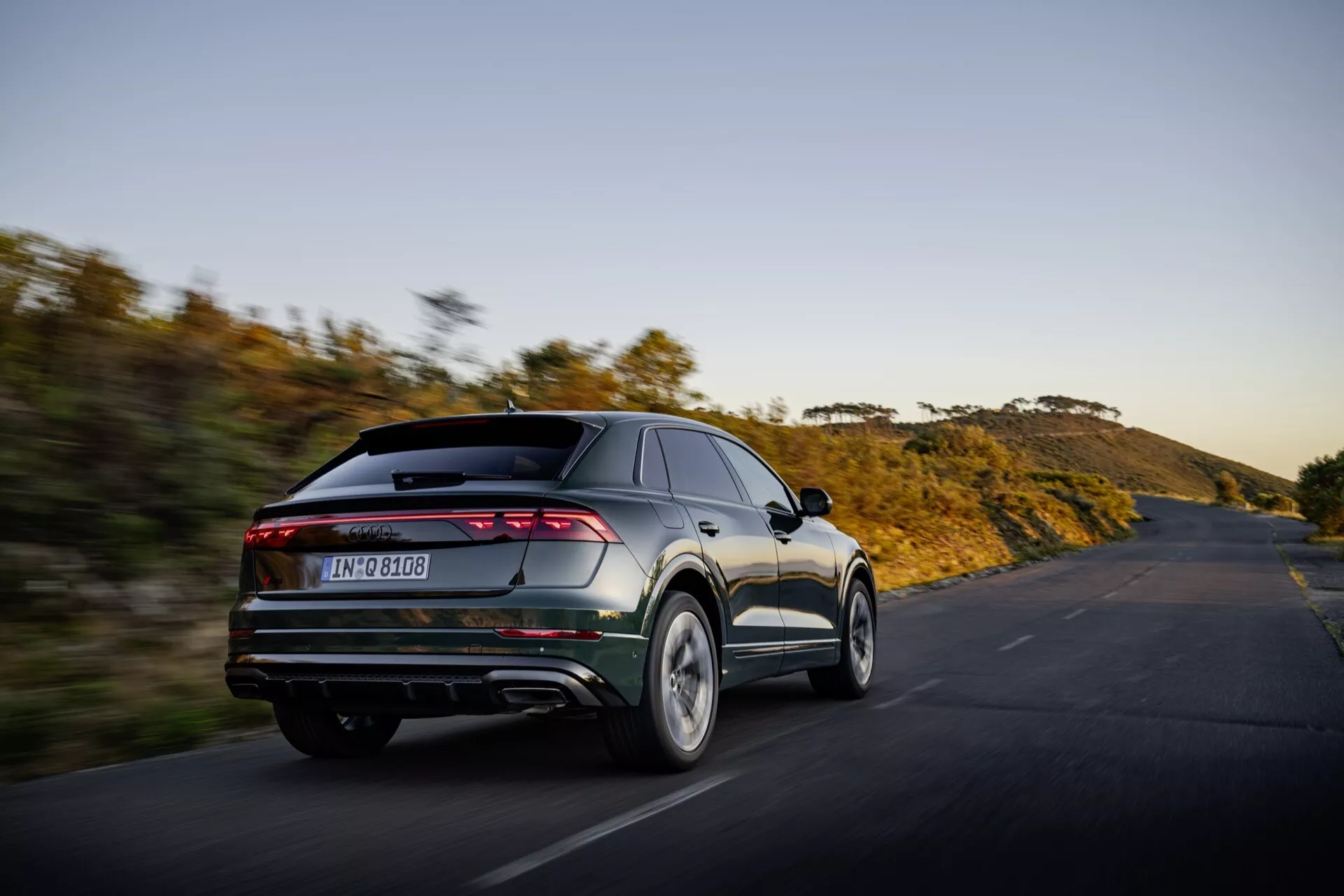 Audi Q8 2023 (4M-Facelift) w ruchu, uchwycone z kąta trzech czwartych z tyłu prezentujące nowoczesne cechy konstrukcyjne pojazdu w tym eleganckie, pełnoszerokościowe poziome tylne światła, podwójne rury wydechowe, tylny dyfuzor i zintegrowany spoiler na tle naturalnego krajobrazu na otwartej drodze o wschodzie lub zachodzie słońca, podkreślając jego orientację na wydajność i zaawansowaną estetykę.