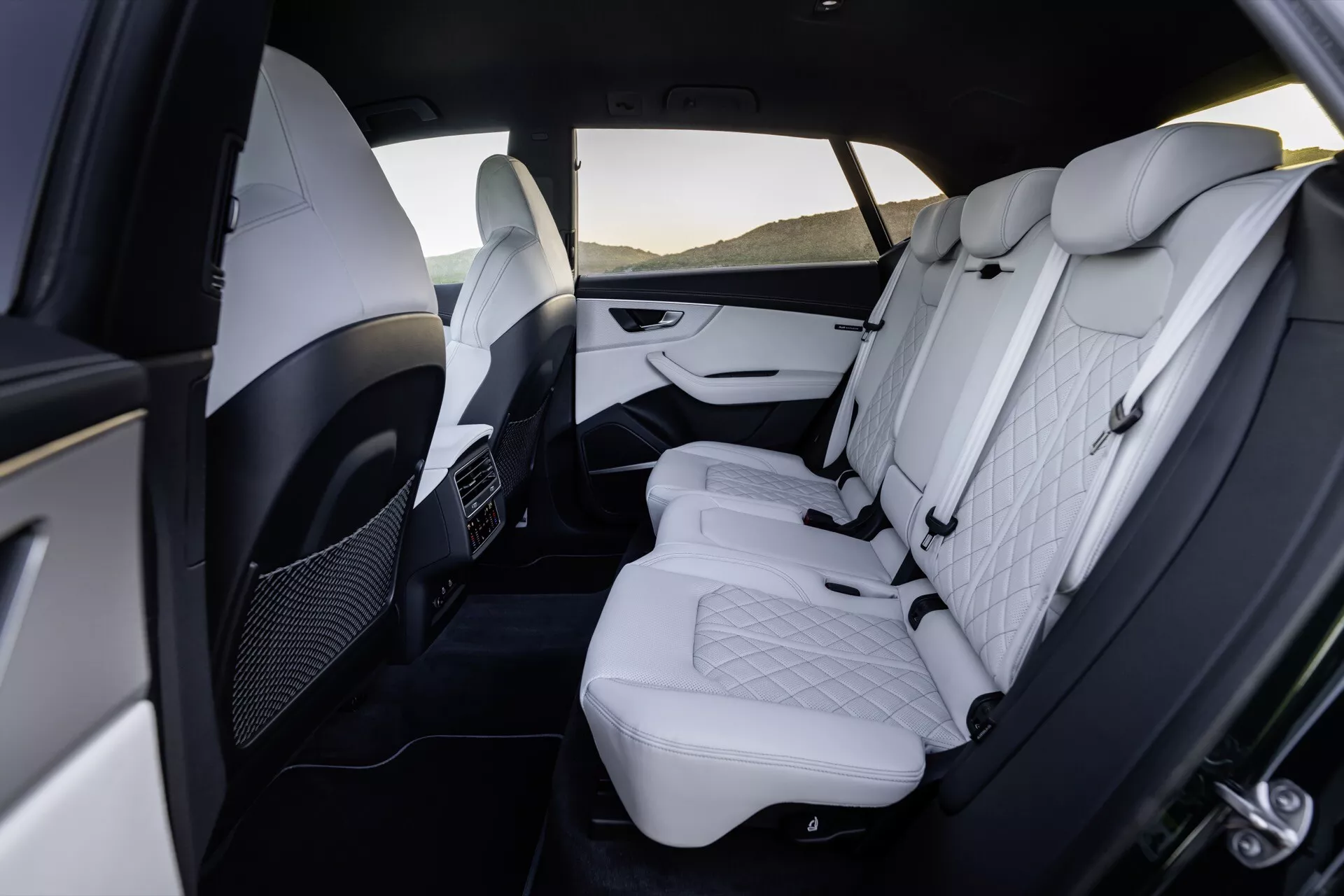 Widok luksusowego wnętrza przedziału tylnej części Audi Q8 2023 (4M Facelift), ukazujący premium białą skórzaną tapicerkę ze wzorem w romby, regulowane zagłówki, dwukolorowe panele drzwiowe z akcentami chromu, składany podłokietnik, klimatyzację dla pasażerów tylnych i przestronne miejsce na nogi.