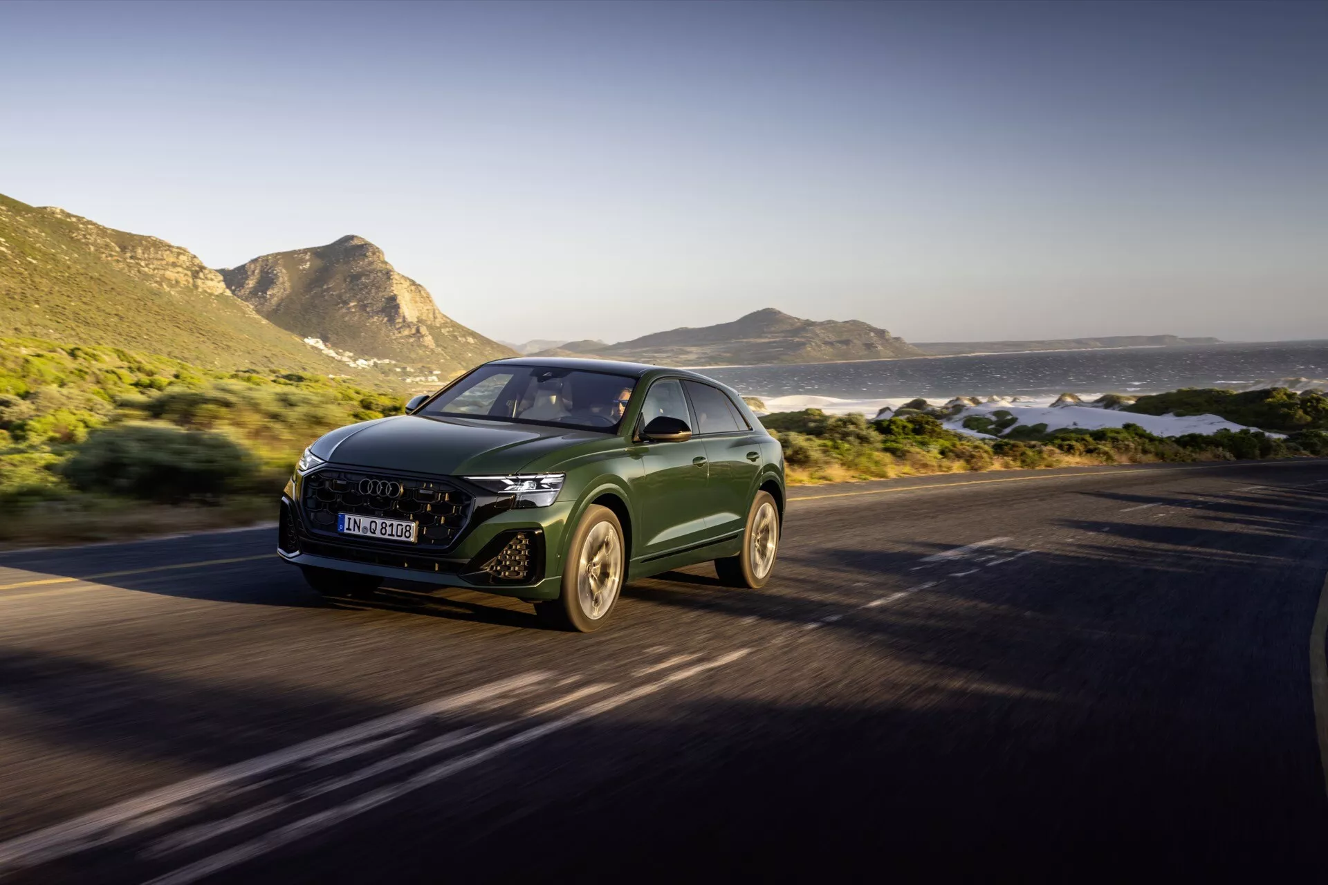 Audi Q8 4M-Facelift 2023 jedzie drogą przybrzeżną, wyróżniające się aerodynamicznym designem, światłami LED i emblematem producenta na grillu, uchwycone z przodu pod kątem trzech czwartych, pokazujące luksusowe SUV w ruchu na malowniczym tle