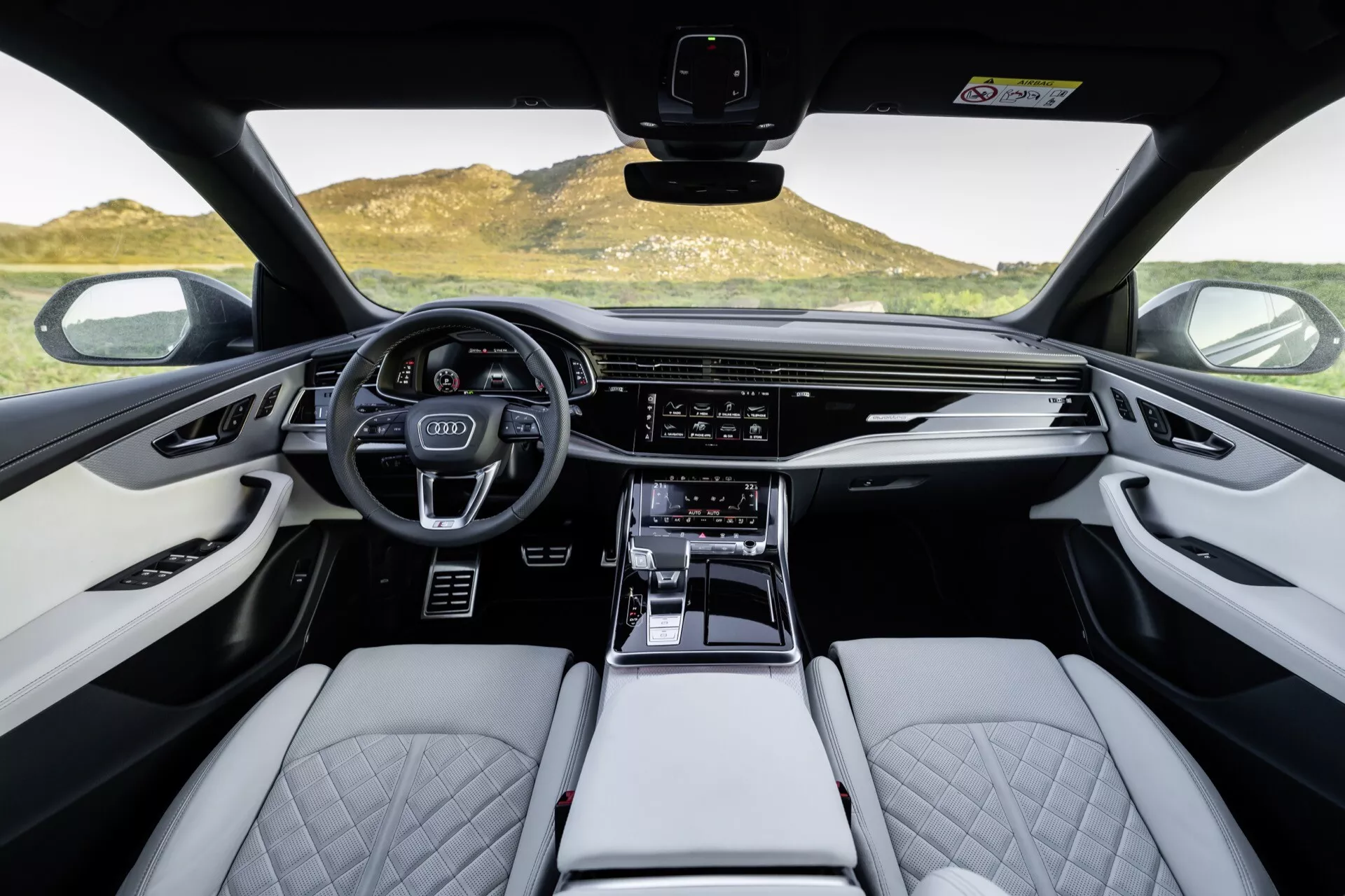 Wnętrze Audi Q8 2023 (4M-Facelift), widok na przednią kabinę z perspektywy pierwszej osoby, z naciskiem na sportową, wielofunkcyjną kierownicę, cyfrowy zestaw wskaźników, szeroki ekran systemu infotainment, jasne skórzane siedzenia z pikowanym wzorem diamentowym i detale wykończenia wysokiej jakości.