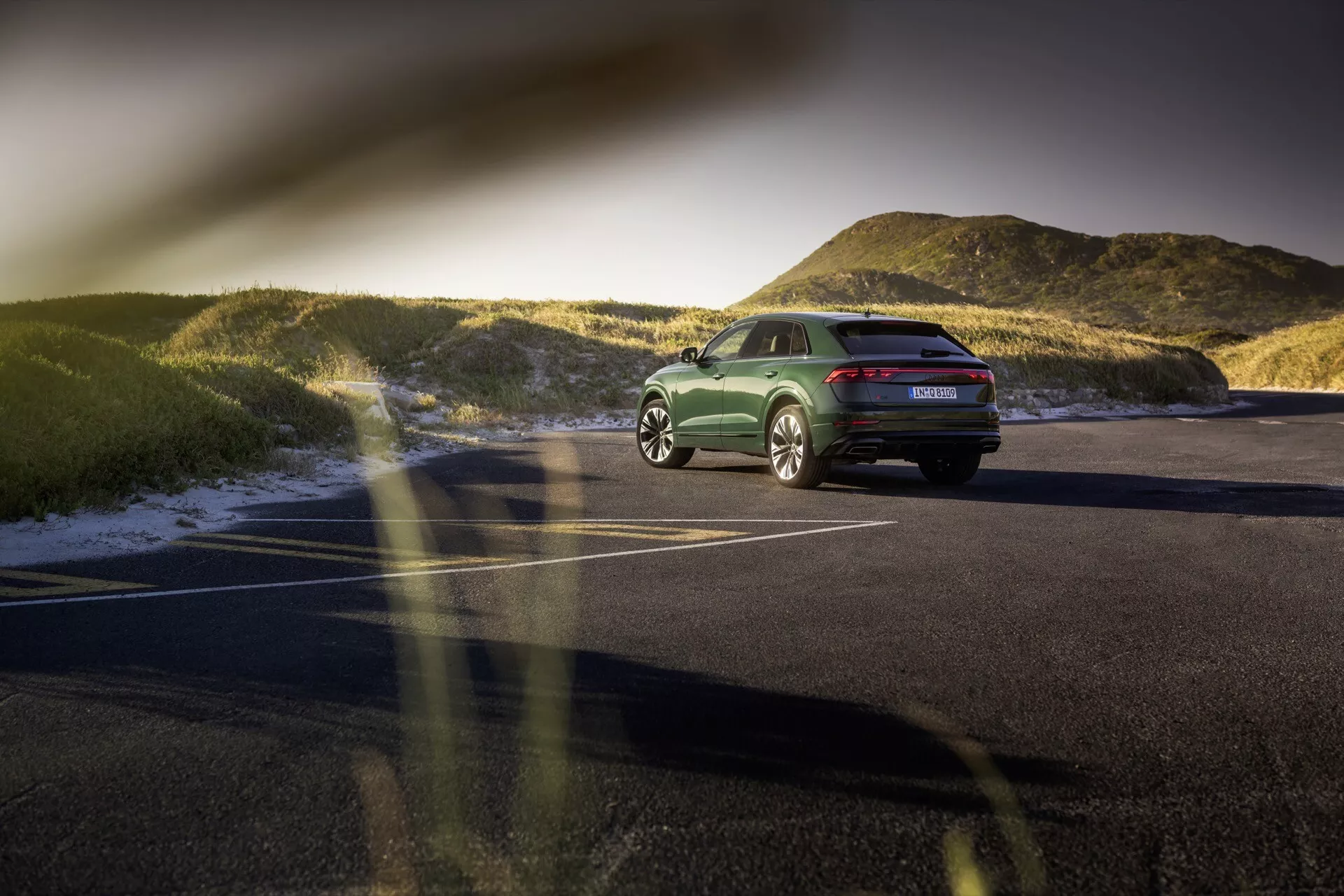 Tylny trzy czwarte widok Audi Q8 (4M-Facelift) 2023 na asfaltowym parkingu o zachodzie słońca, prezentujący jego postawę, linie projektowe, tylne światła LED, koła ze stopu i polerowaną zieloną wykończeniem z naturalnym krajobrazem w tle.