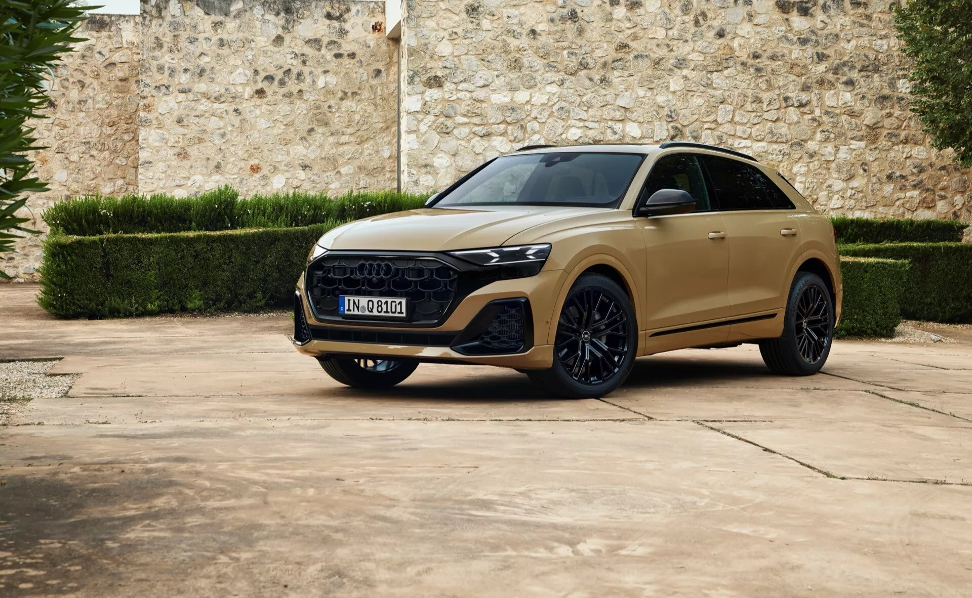 Luksusowy crossover SUV Audi Q8 2023 w widoku trzy czwarte z przodu zaparkowany na brukowanej nawierzchni z tłem starego kamiennego muru, prezentujący eleganckie reflektory LED, wyrazistą sześciokątną atrapę chłodnicy, wyrzeźbione linie maski, duże koła ze stopu i możliwy panoramiczny dach.