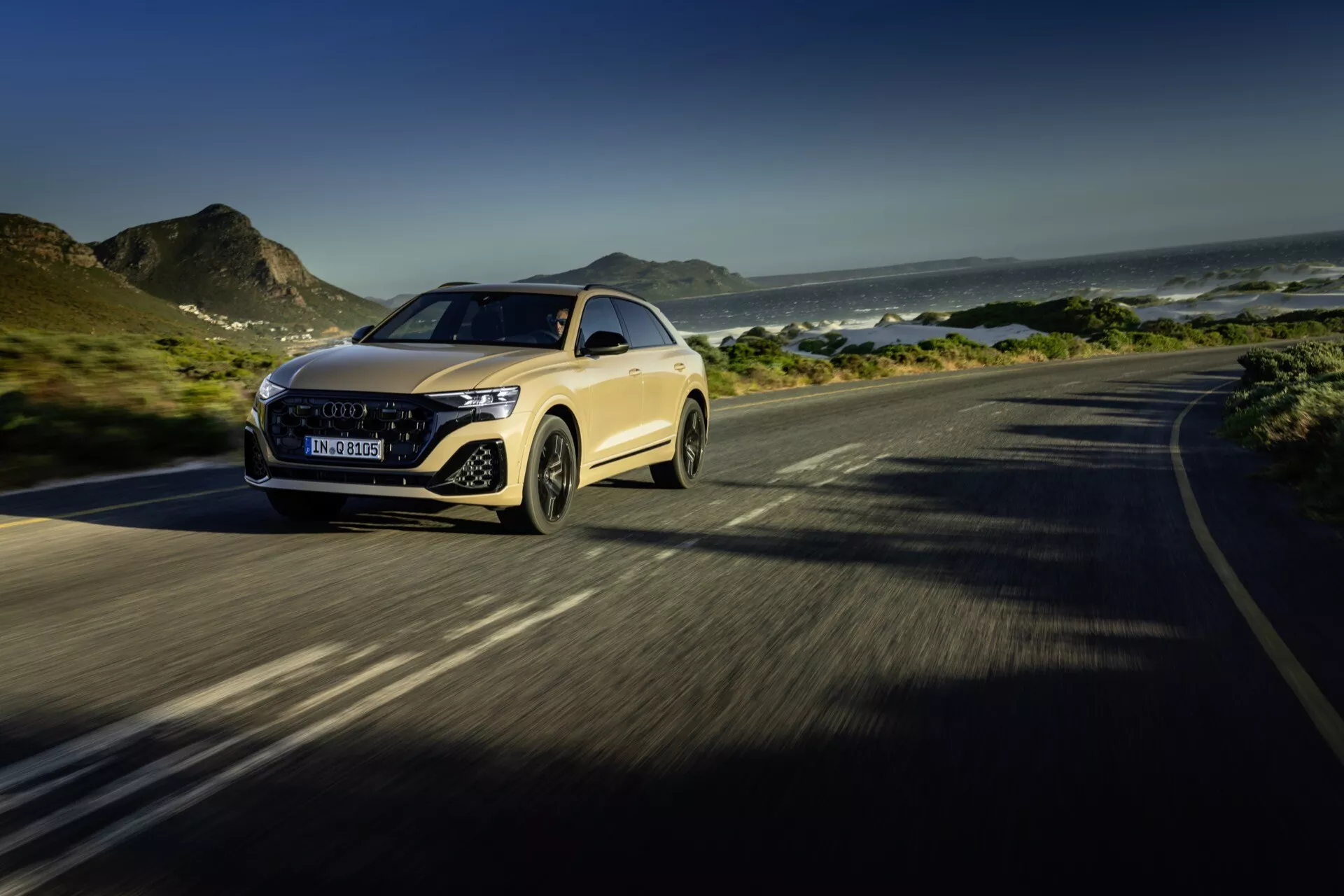 Audi Q8 4M facelift 2023 w ruchu na nadmorskiej drodze, podkreślające zaawansowane reflektory LED luksusowego samochodu, wyrazistą atrapę, aerodynamiczną maskę i dynamiczne prowadzenie, z tłem gór i oceanu, z naciskiem na cechy konstrukcyjne i technologiczne.