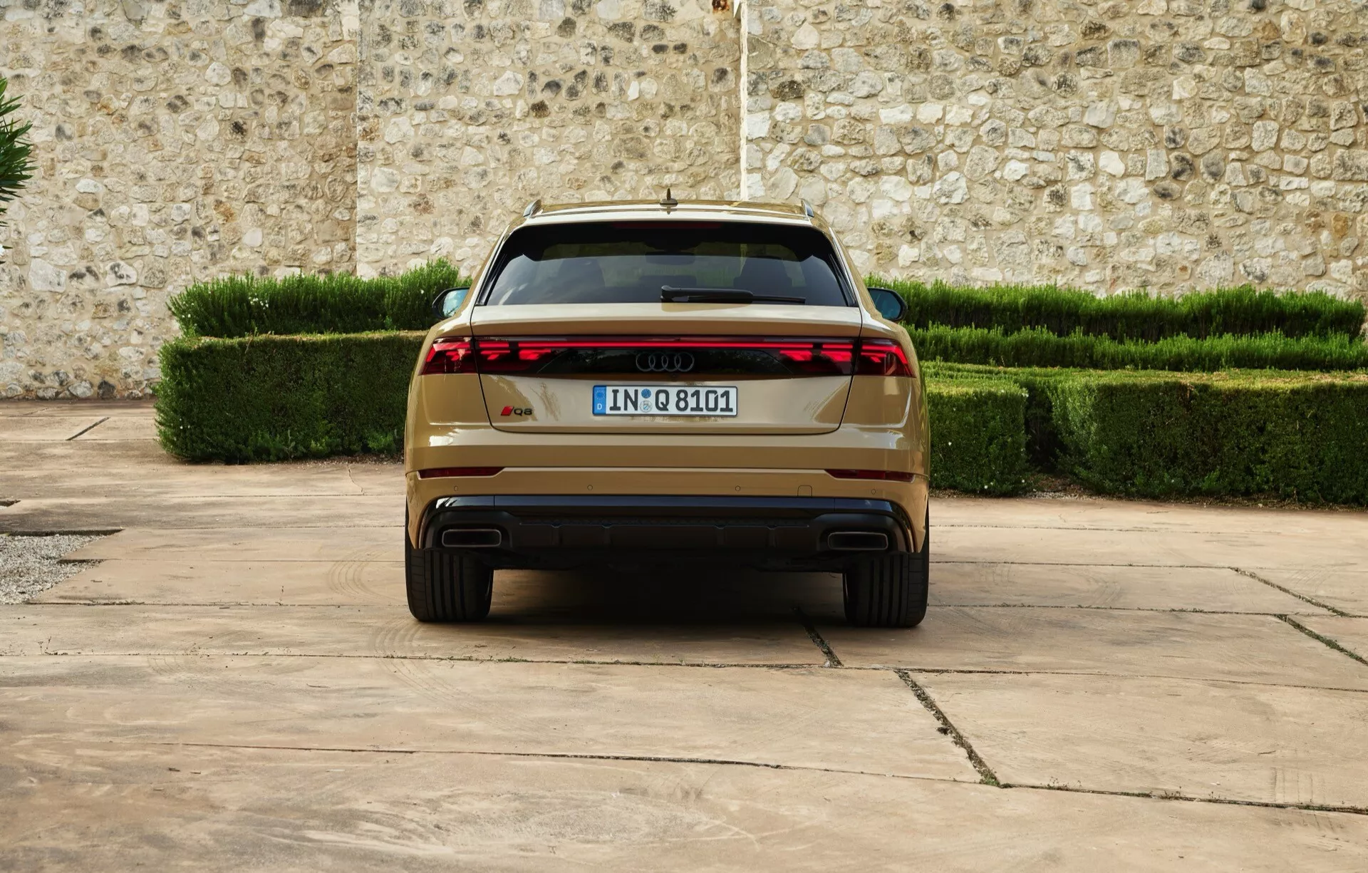 Audi Q8 2023 (4M-facelifting) prezentujące tylny widok z nowoczesnymi, LEDowymi światłami tylnymi, podwójnymi wydechami, europejską tablicą rejestracyjną, zaparkowane na utwardzonej powierzchni na tle kamiennego muru i przyciętych zielonych żywopłotów, podkreślające luksusowe i zaawansowane funkcje designu